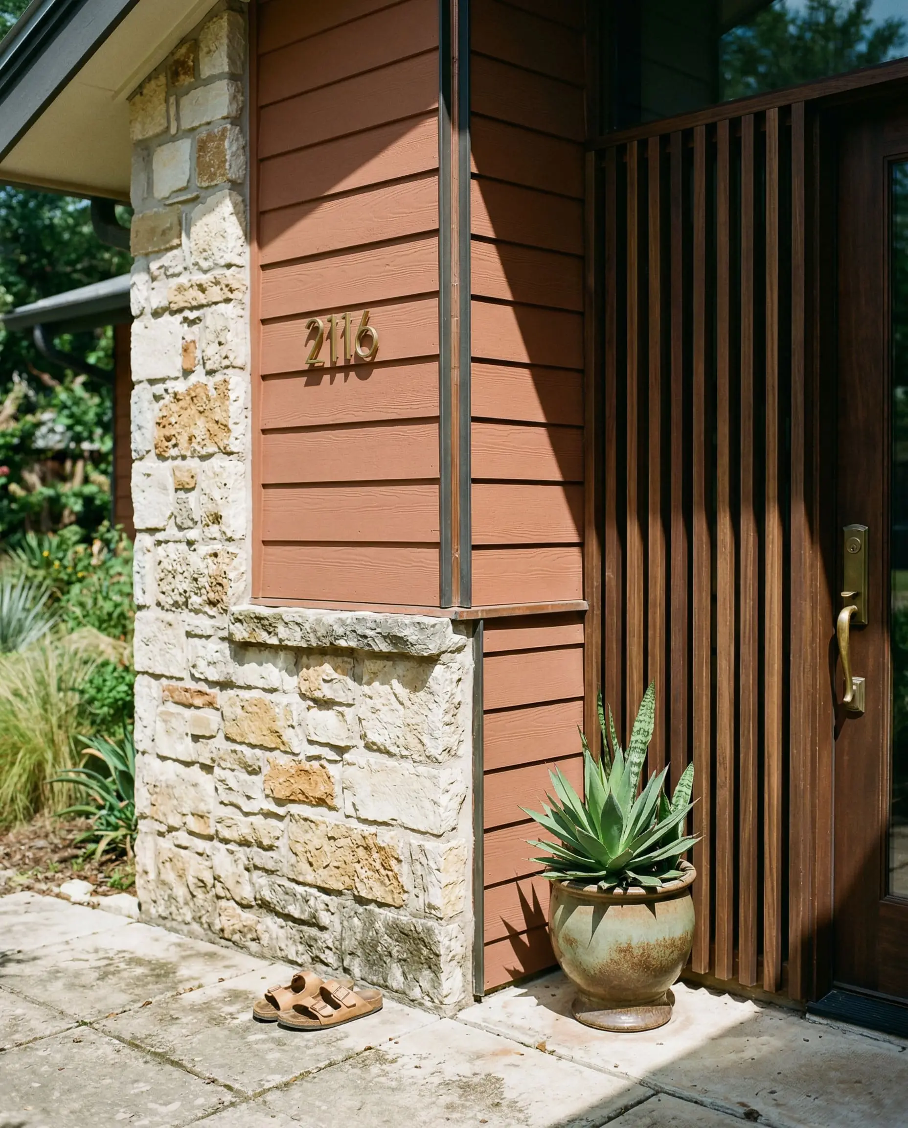 Bright natural sunlight casts sharp shadows across the intersection of earthy rust painted siding, natural stone masonry, and a dark walnut wood slat screen.