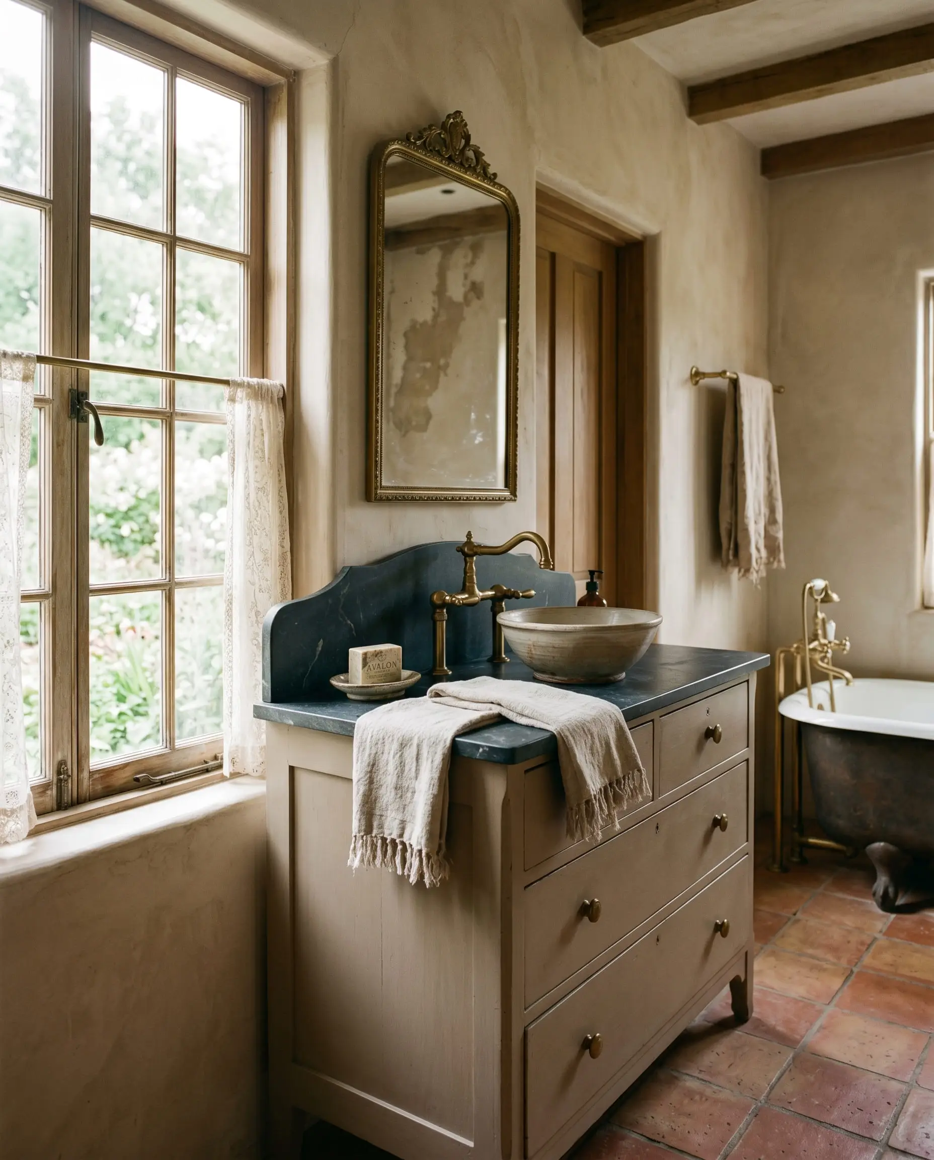 A repurposed antique dresser vanity painted in a historical taupe features unlacquered brass fixtures, a honed soapstone counter, and a folded linen towel.