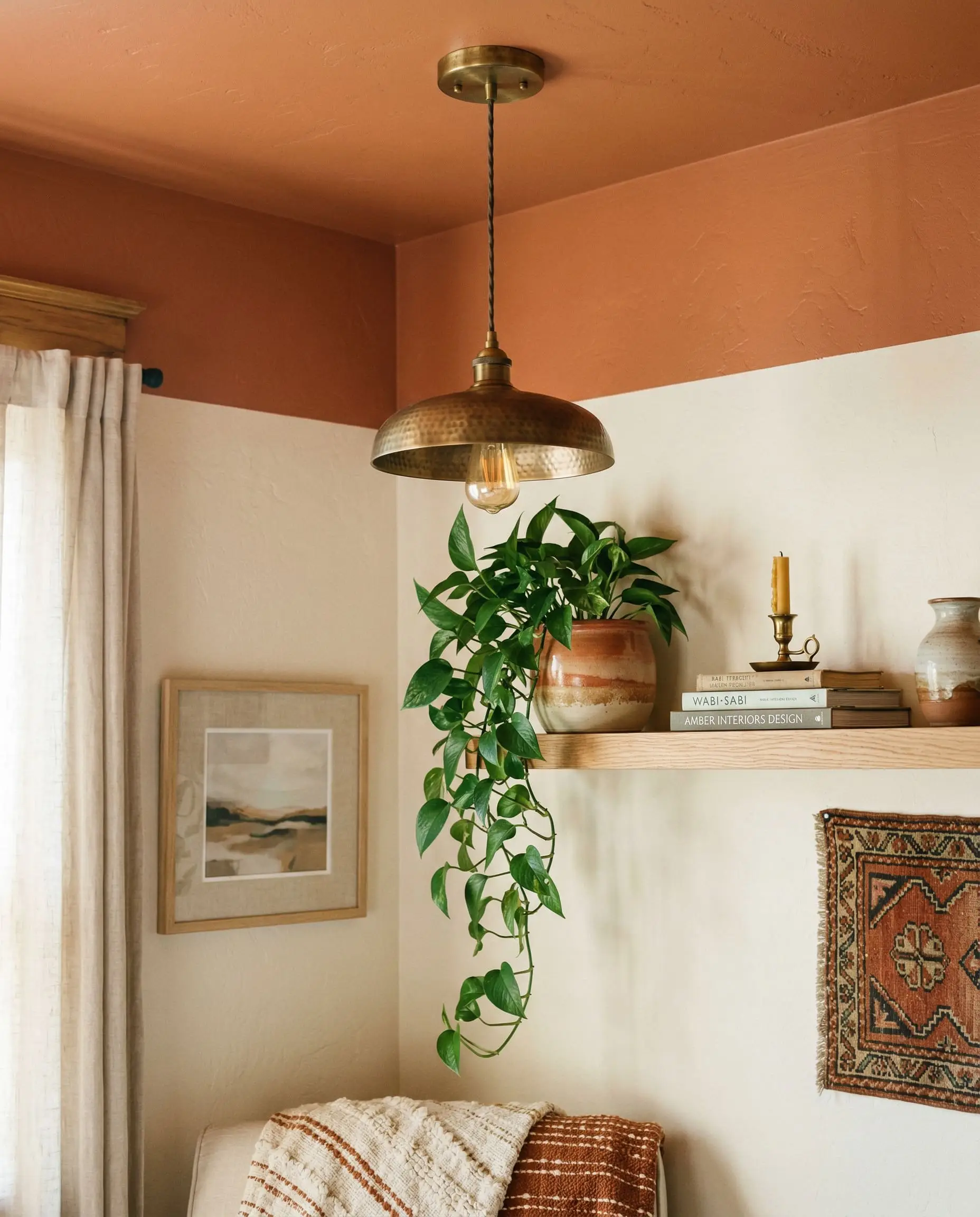 A hanging unlacquered brass pendant and a trailing indoor plant sit near a bedroom corner where a warm clay upper wall meets crisp white lower walls.