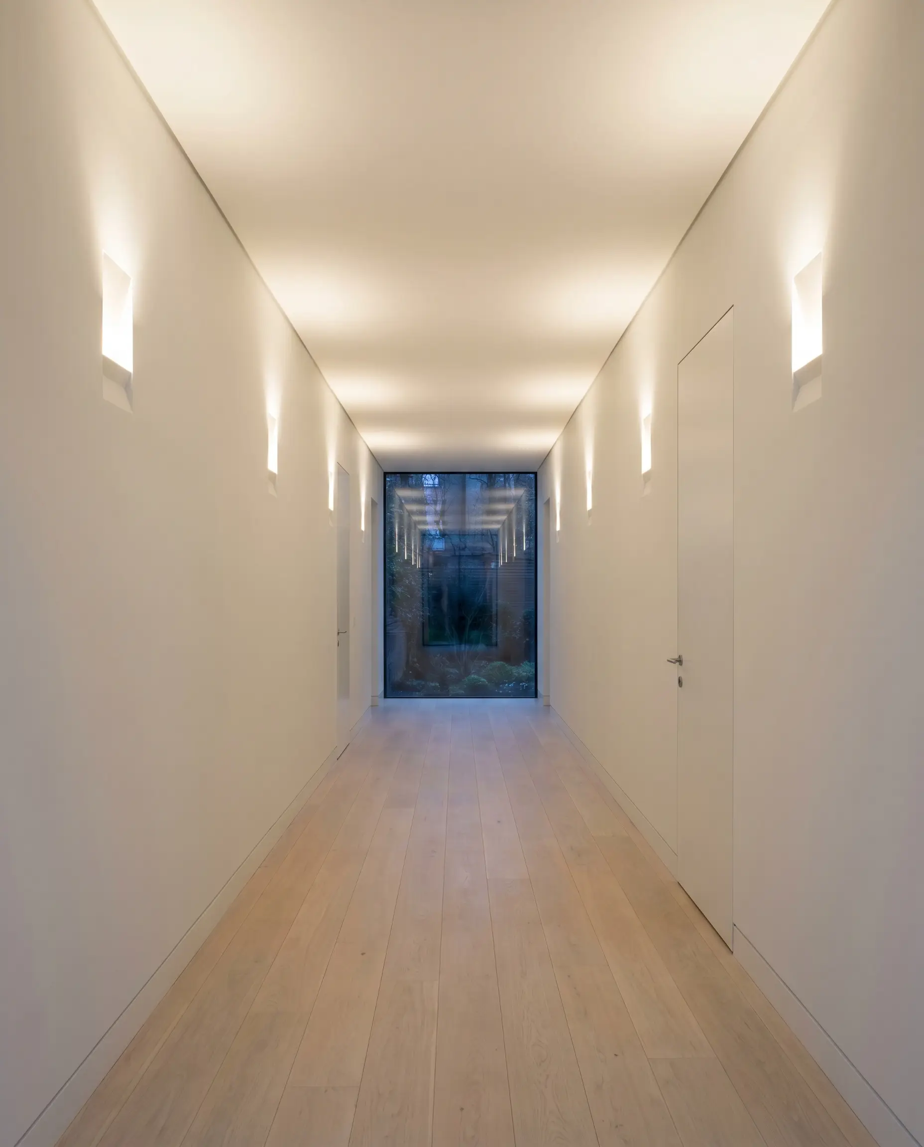 Architectural wall sconces cast a warm 3000K glow up and down a narrow windowless corridor featuring a high-reflectance base wall finish.