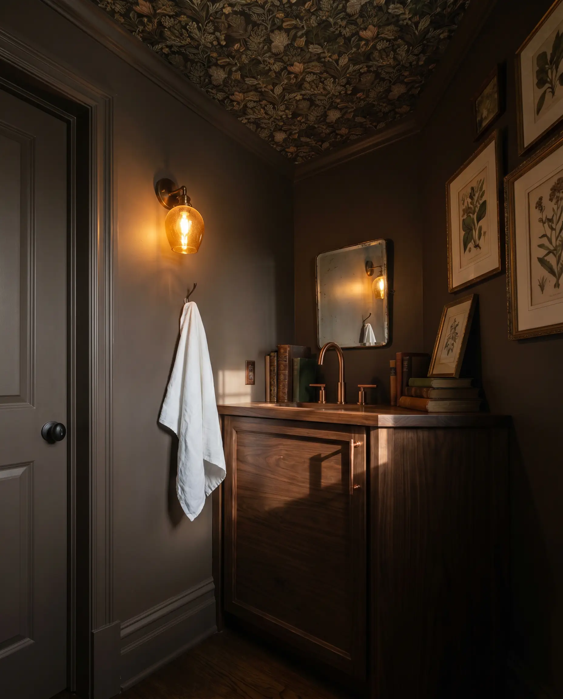 A glowing amber glass sconce illuminates heavily patterned botanical ceiling wallpaper, chalky brown-gray walls, a polished copper faucet, and a dark walnut vanity.