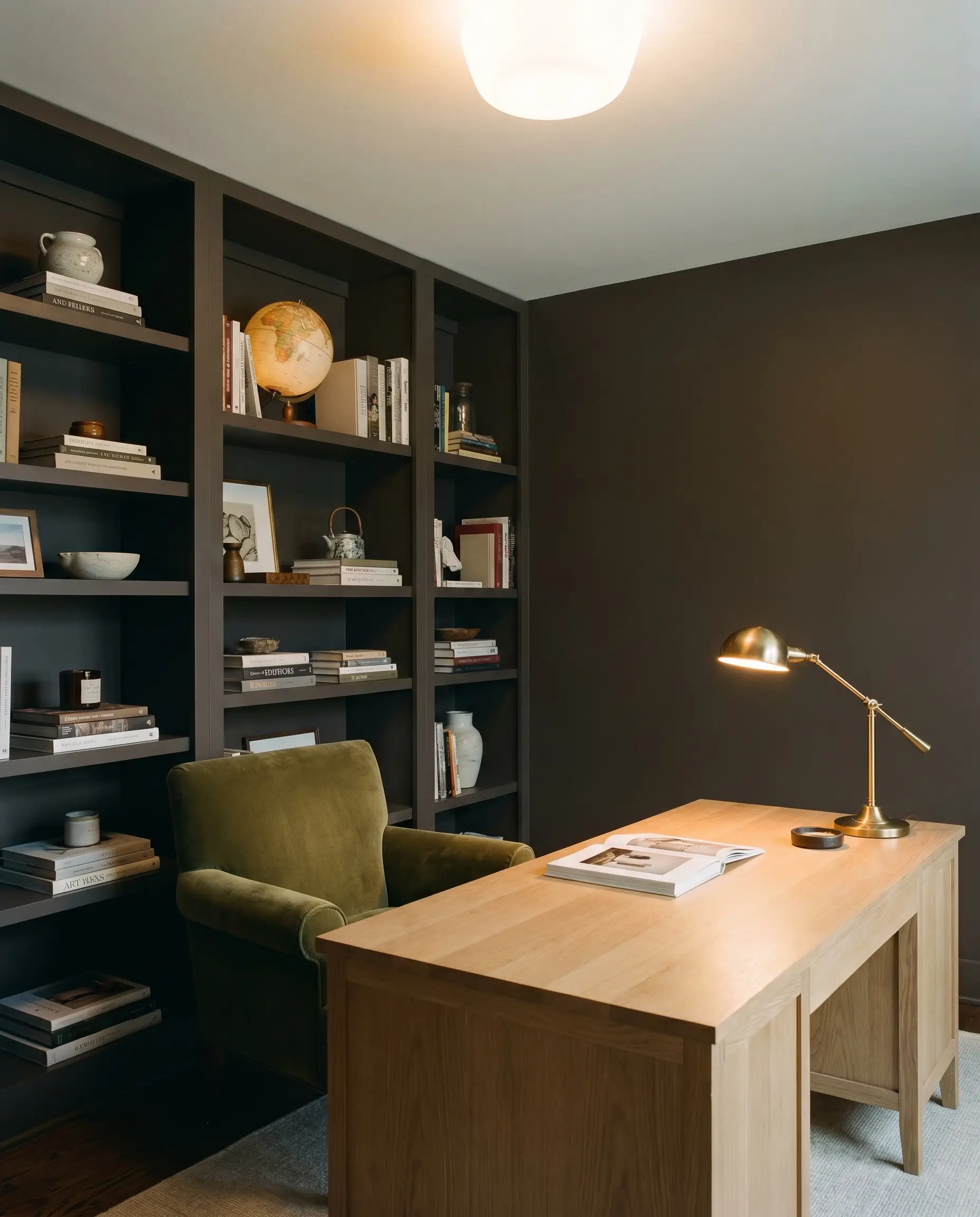 Floor-to-ceiling built-in bookcases in a grounding neutral dark brown surround a rich white oak desk and a heavy olive velvet reading chair.