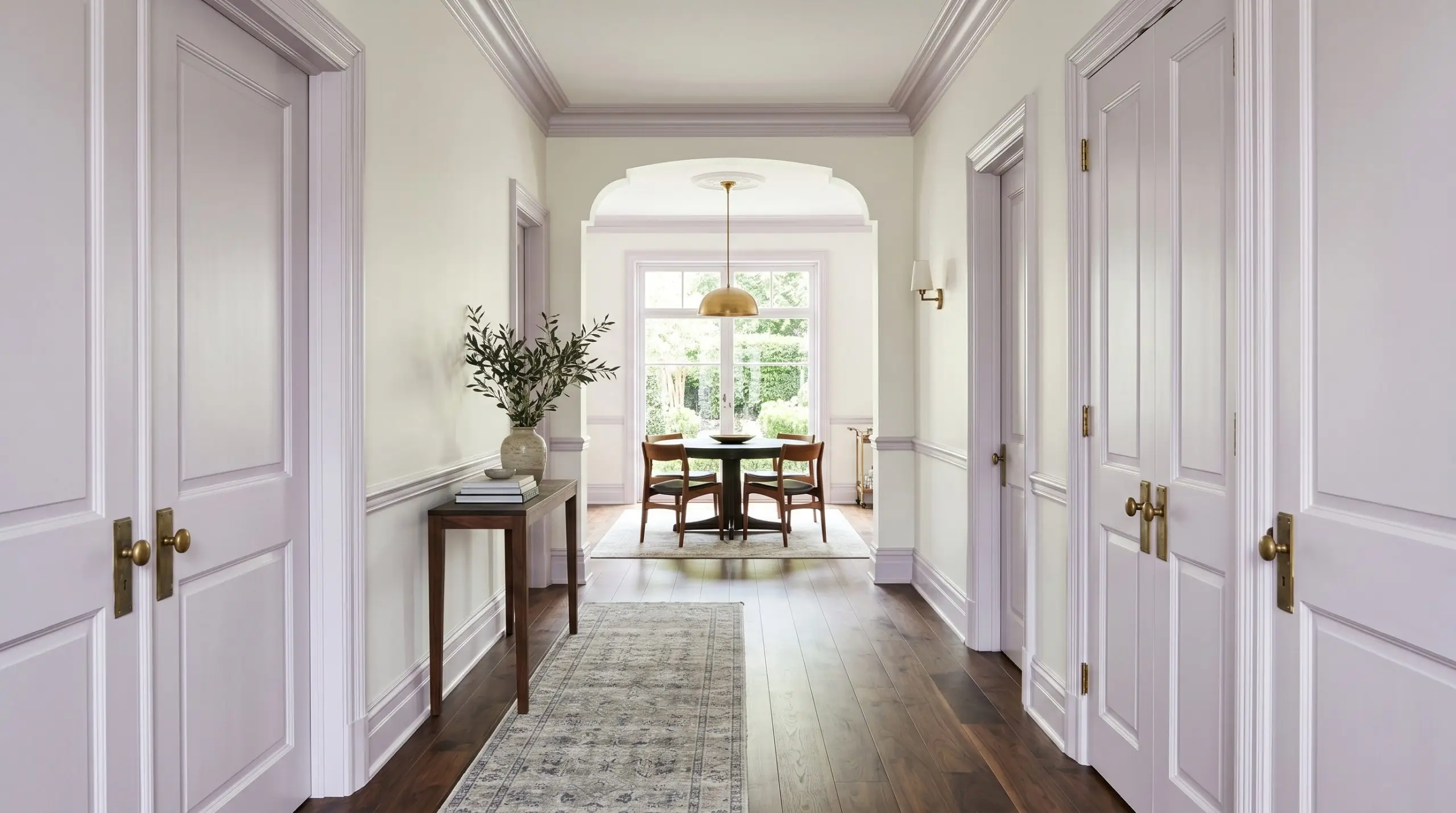 Unlacquered brass hardware accents soft violet interior doors and crown molding set against warm white walls and dark walnut flooring in a tailored hallway.