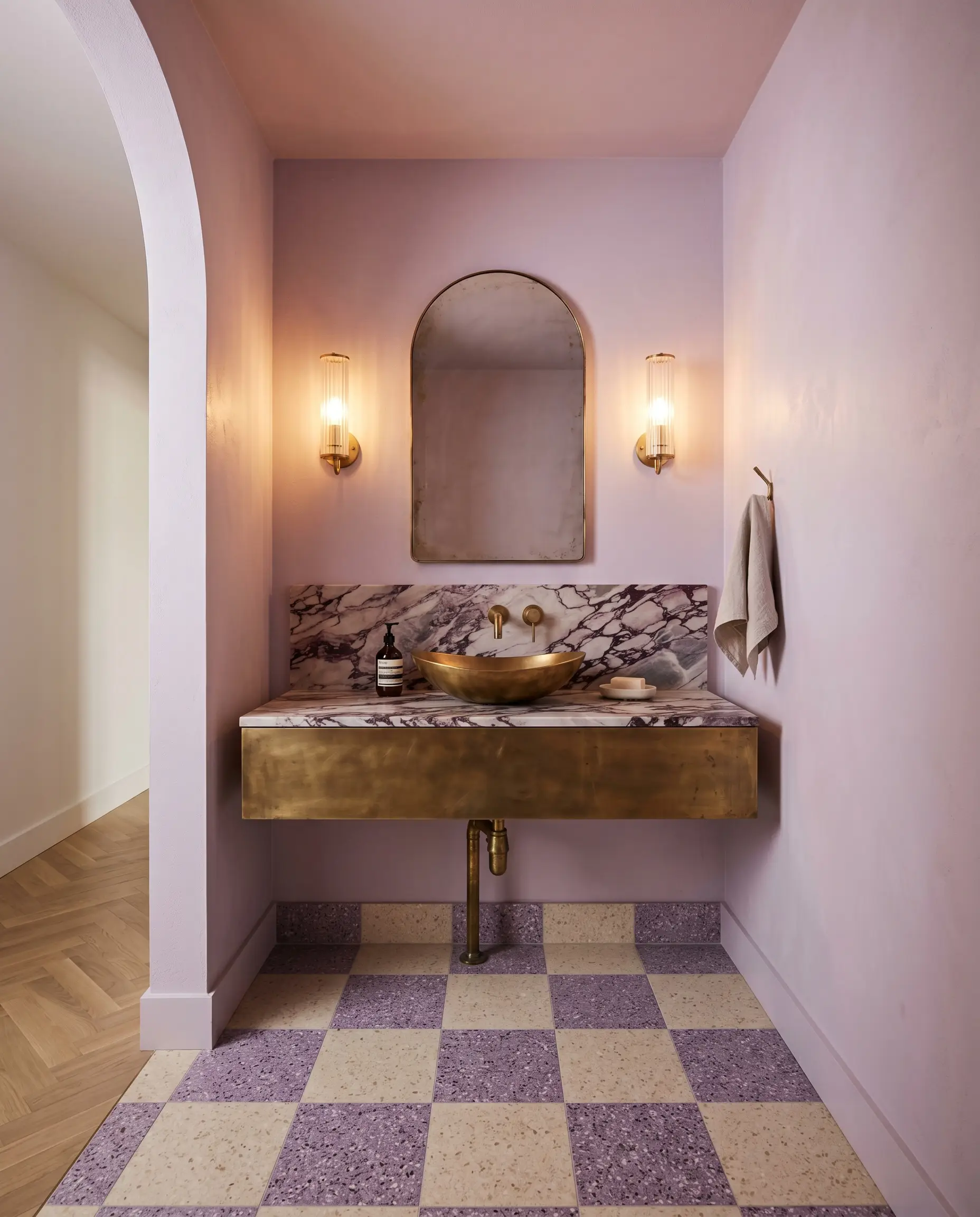 An unlacquered brass floating vanity and heavily veined honed marble countertop highlight a jewel box powder room entirely color-drenched in Benjamin Moore Misty Lilac (Hex #EAE3F0).