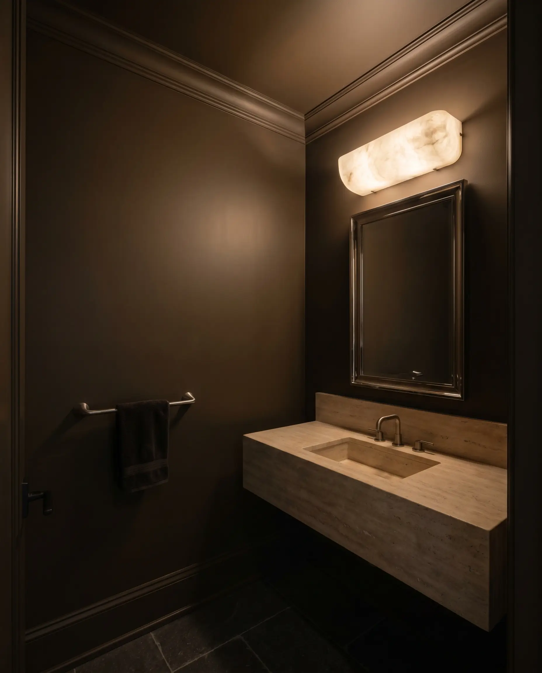 A travertine stone vanity and polished nickel mirror anchor a windowless powder room completely color-drenched in Benjamin Moore Night Horizon (Hex #4B433D).