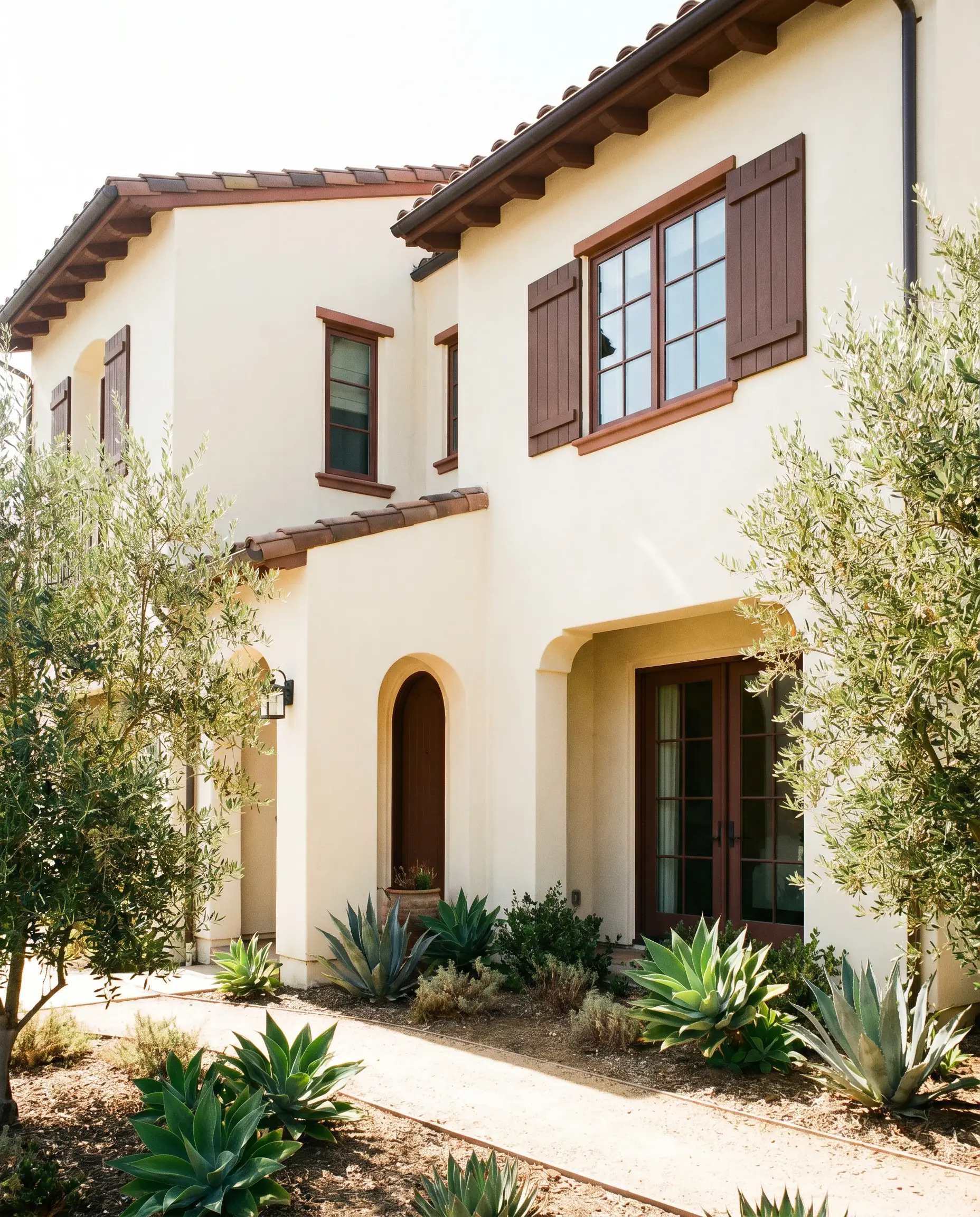 A creamy stucco Spanish Revival exterior facade features natural landscaping alongside trim, shutters, and fascia painted in the earthy neutral tone of Clove.