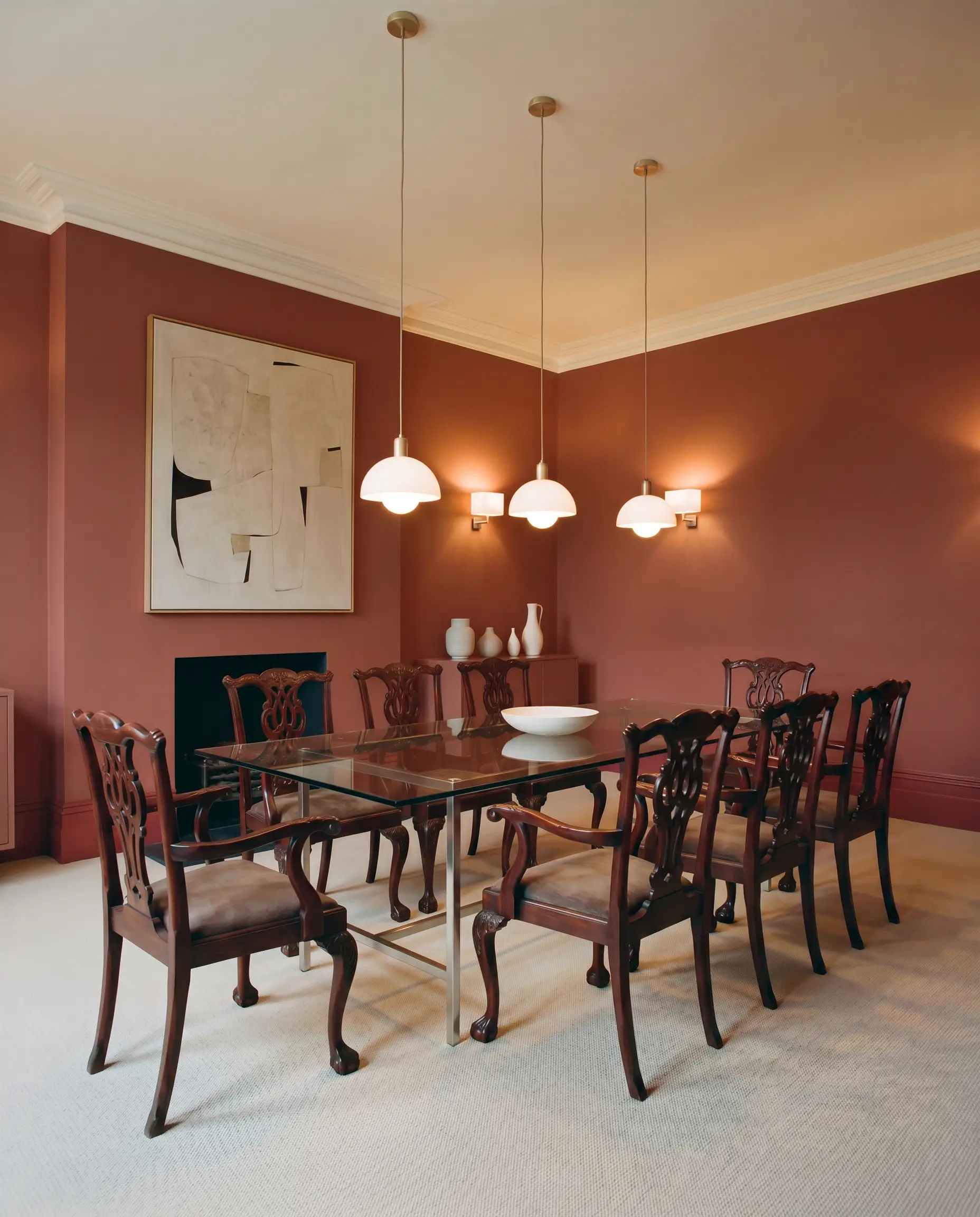 A formal dining room features rich heritage red walls and baseboards surrounding a sleek Bauhaus-inspired glass table and heavily carved mahogany chairs.
