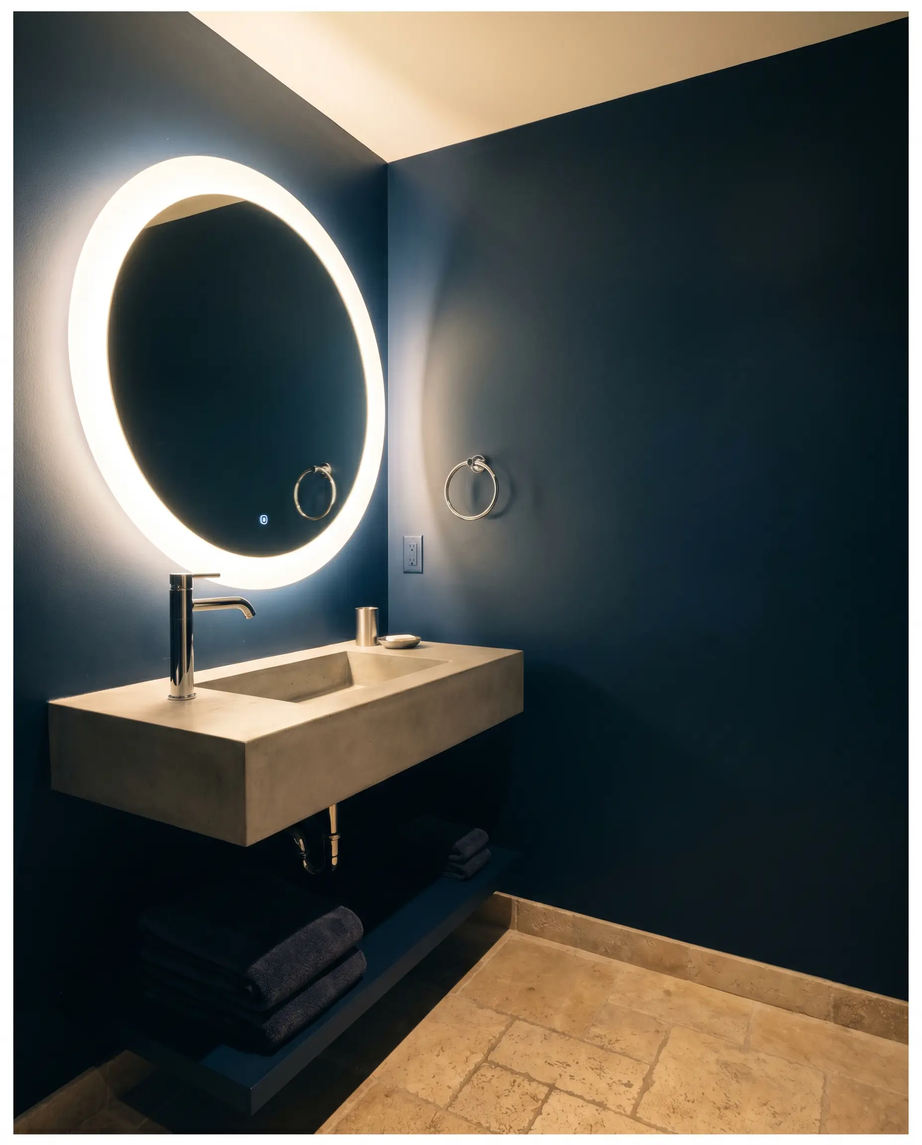 A windowless jewel-box powder room pairs tumbled limestone flooring with rich walls painted in Behr Midnight Blue (Hex #4c565e) and polished chrome hardware.
