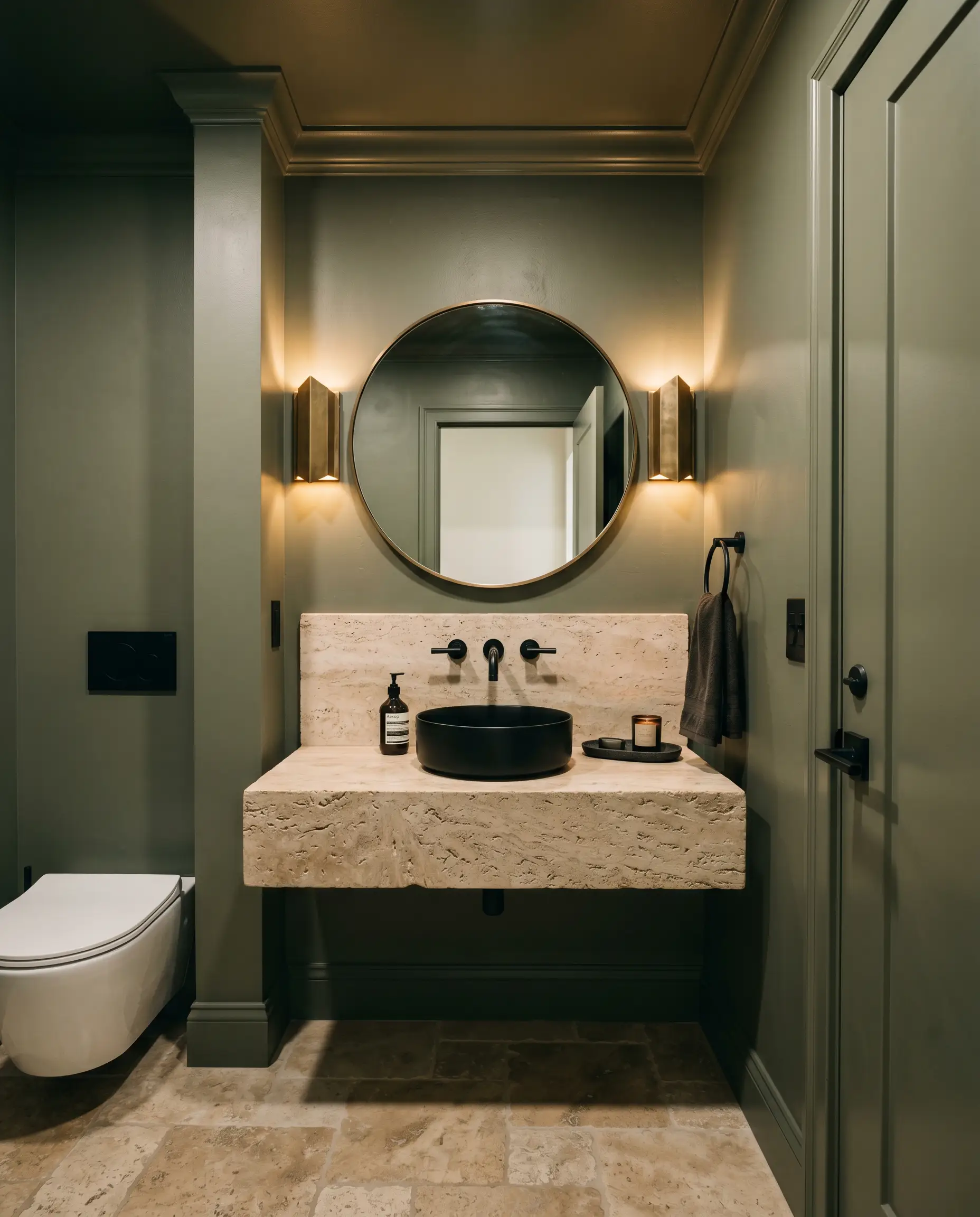 A tumbled travertine vanity and a highly reflective mirror define this jewel-box powder room, surrounded by muddy sage walls, trim, and matte black iron hardware.