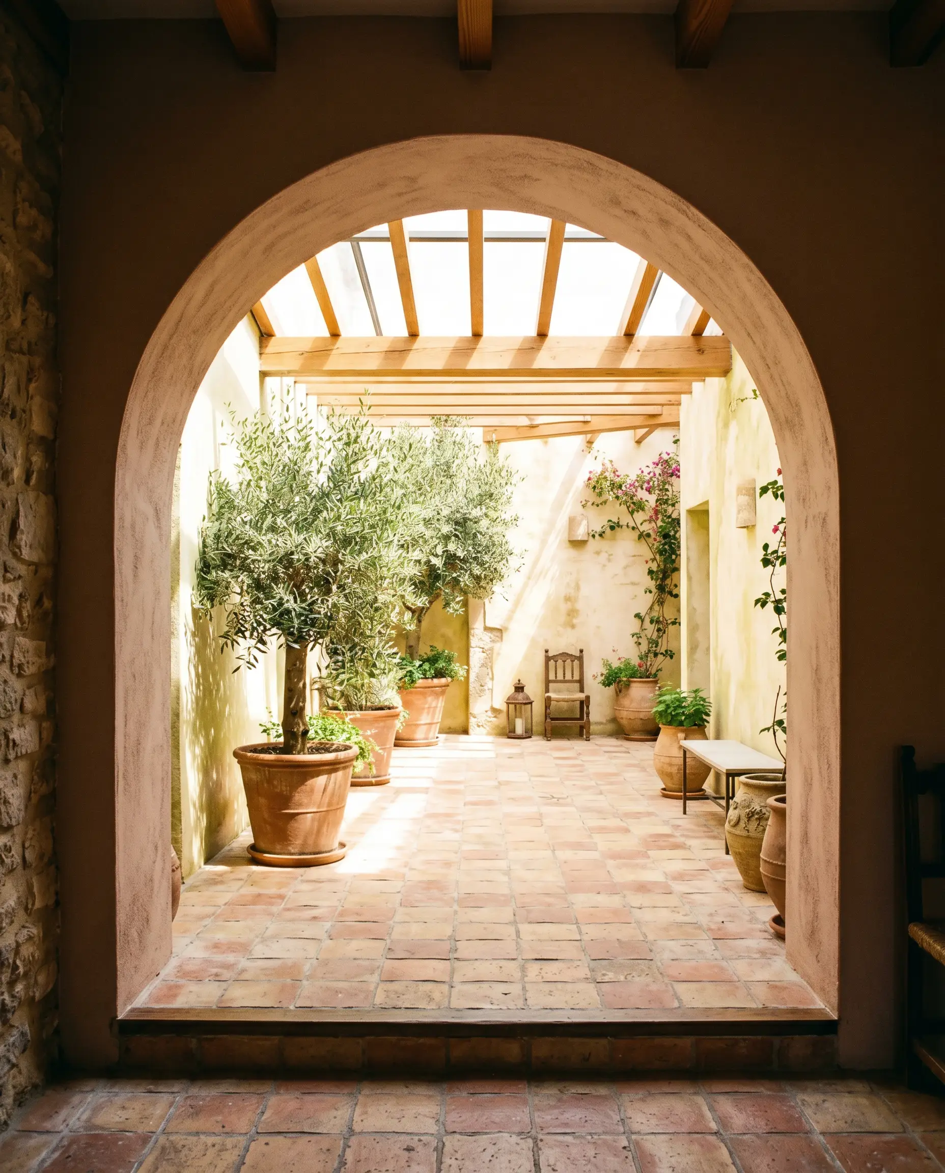 Tumbled terracotta floor tiles and lush, oversized potted olive trees fill a sun-drenched enclosed patio framed by a large, earthy beige architectural archway.