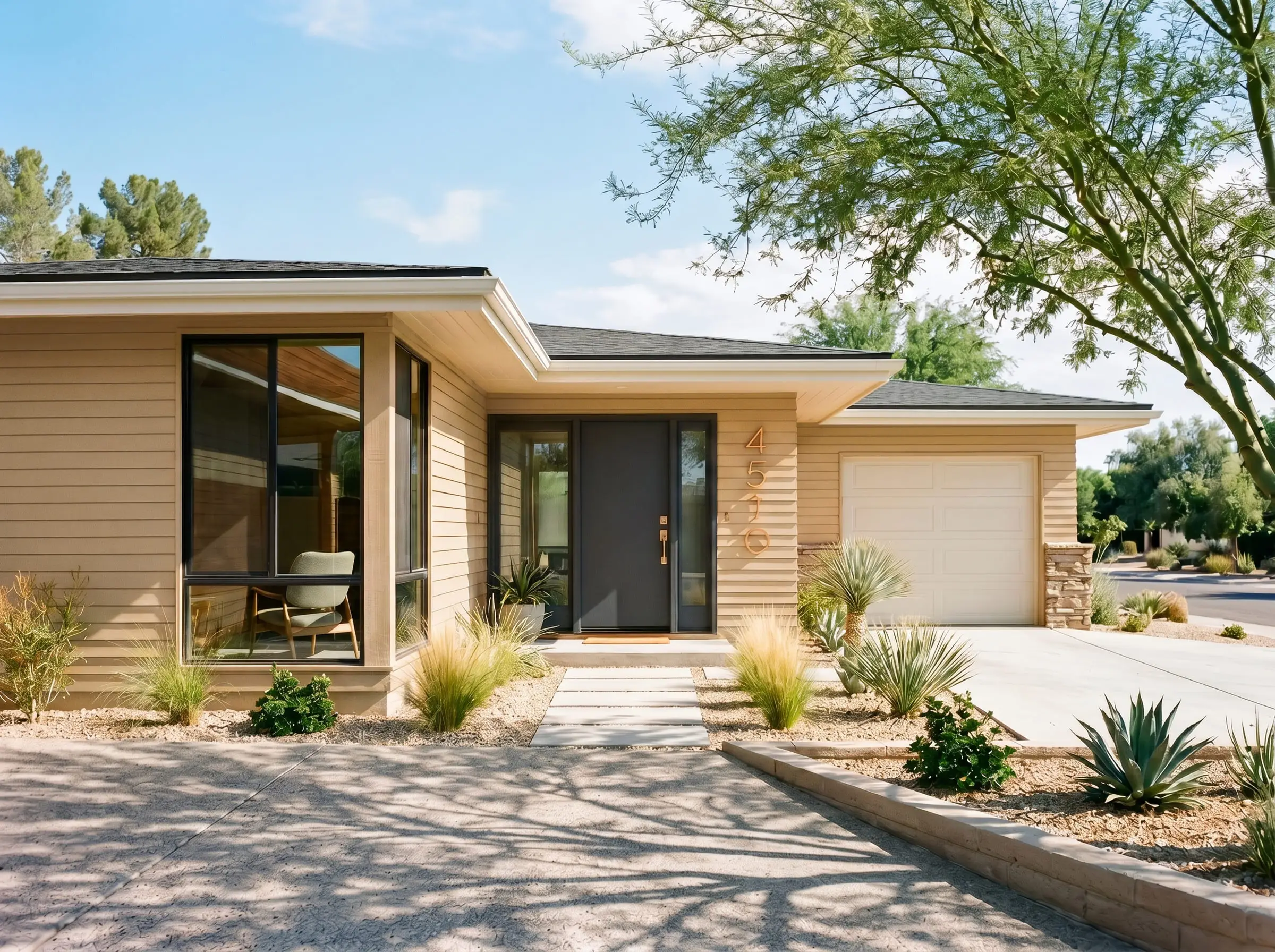 A mid-century ranch home exterior features warm khaki tone siding, alabaster eaves, a bold charcoal gray front door, and oversized modern brushed copper house numbers.