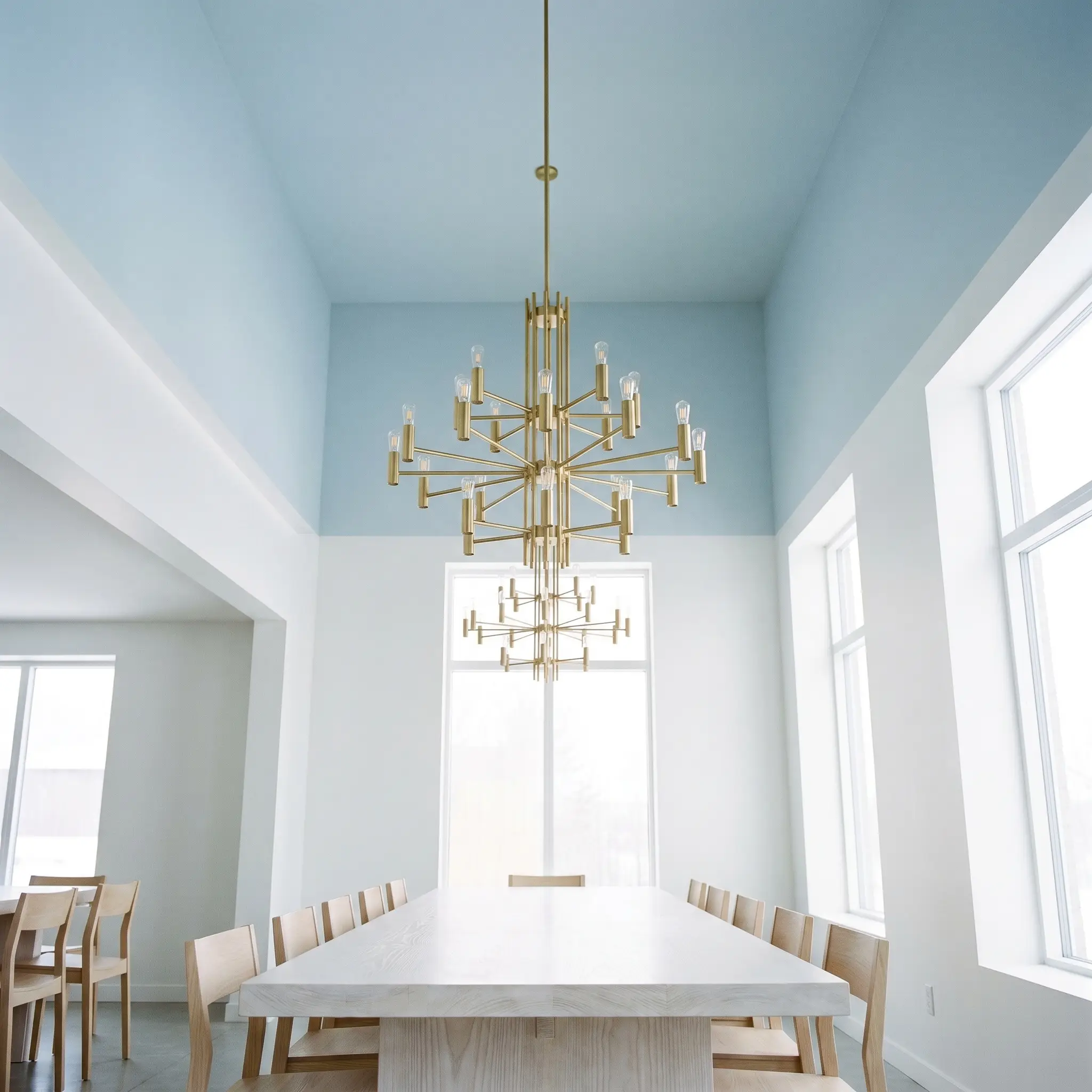 A modern brass chandelier hangs from a pastel blue painted ceiling over a whitewashed ash table surrounded by crisp gallery white walls.