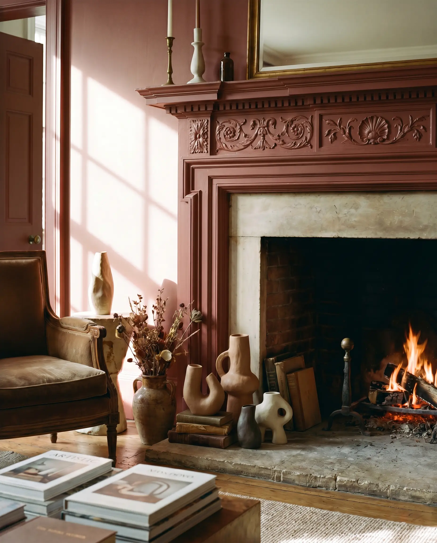 An ornate, carved wooden mantelpiece painted in a rich heritage red frames a living room fireplace, with modern asymmetrical ceramics styled on the hearth.