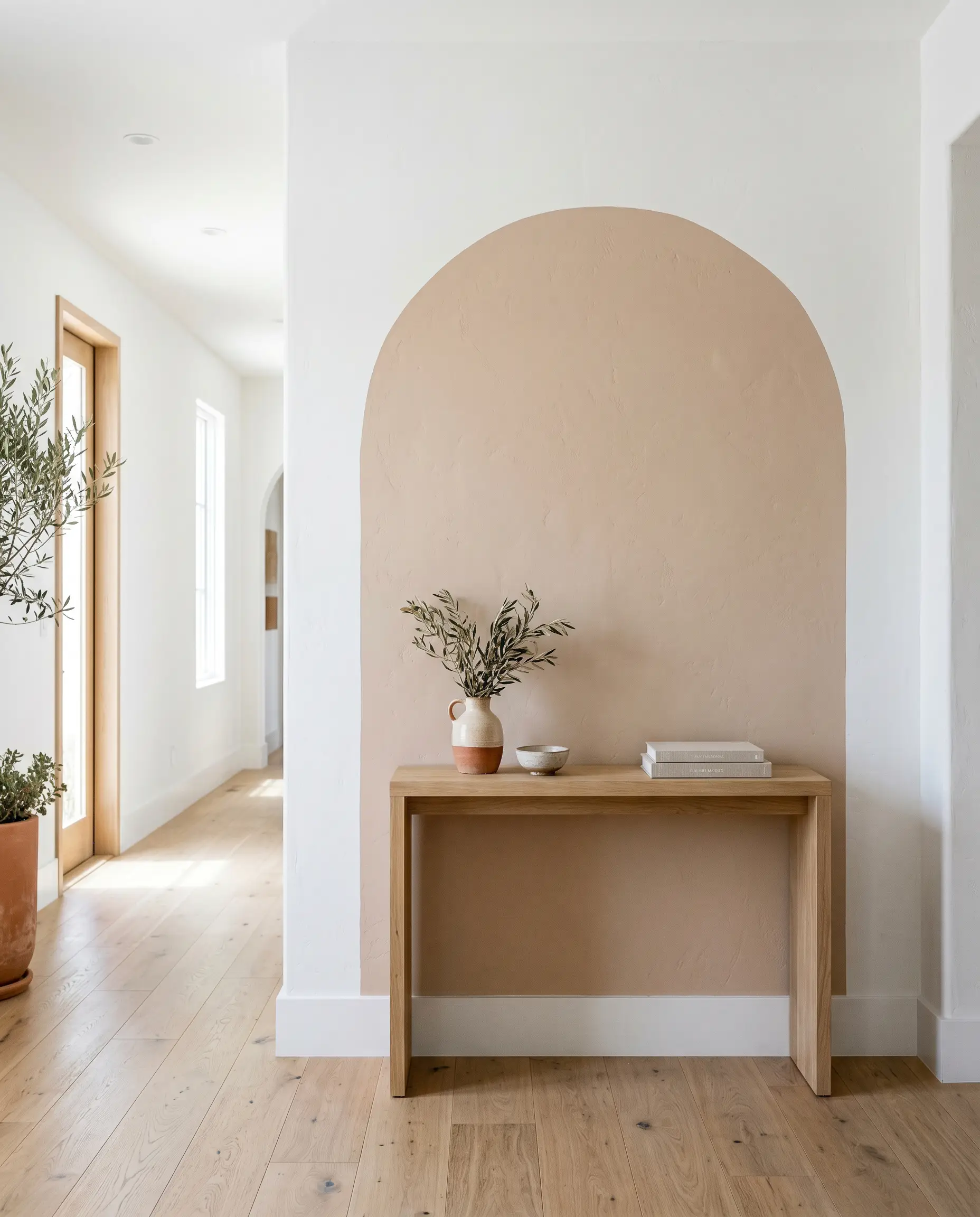 A small wooden console table sits on white oak flooring centered beneath a bold oversized Mediterranean archway painted in Artisan Tea (Hex #dac2af).