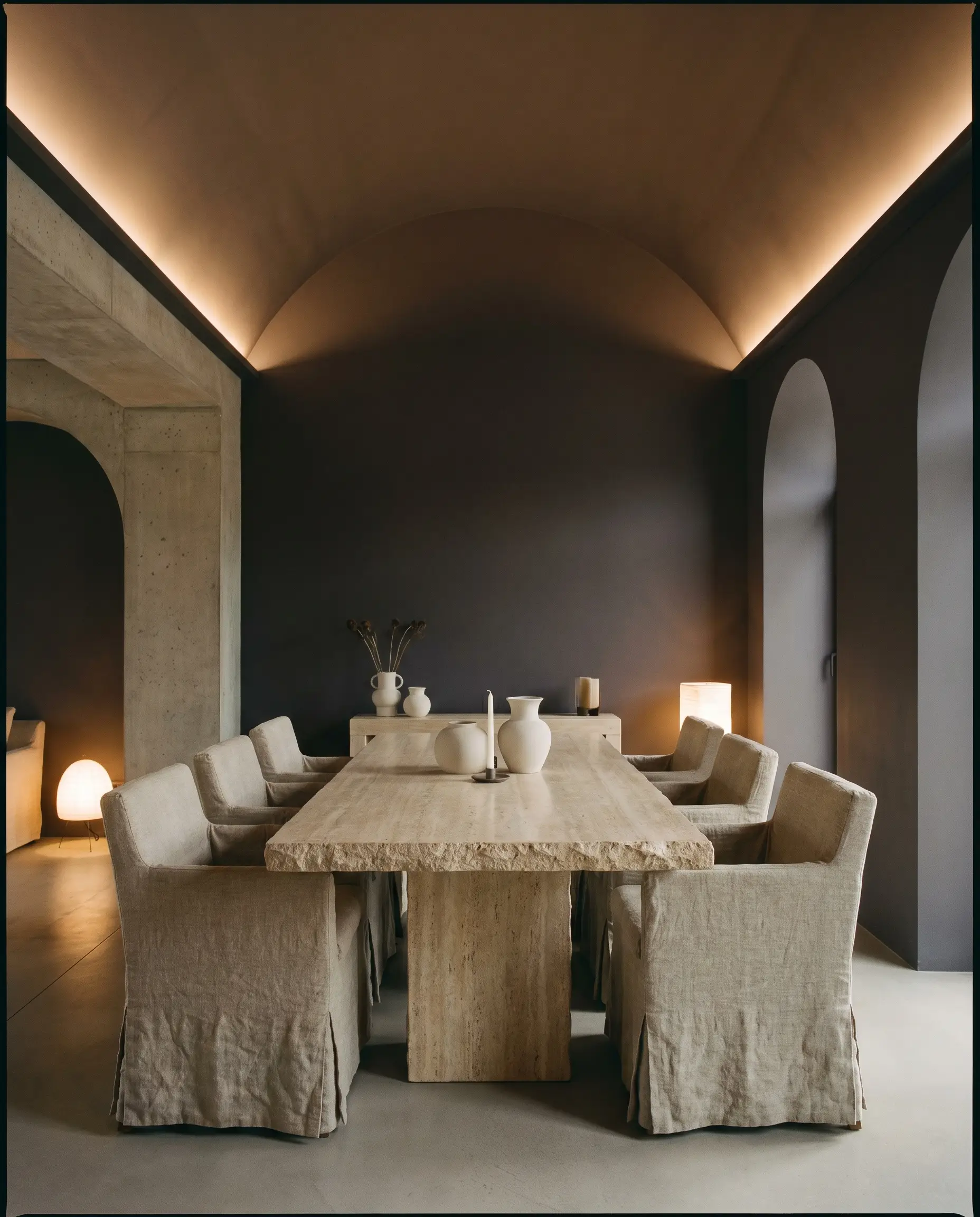 An intimate dining room seamlessly color-drenched in a red-based black features a travertine dining table surrounded by slub linen slipcovered chairs.