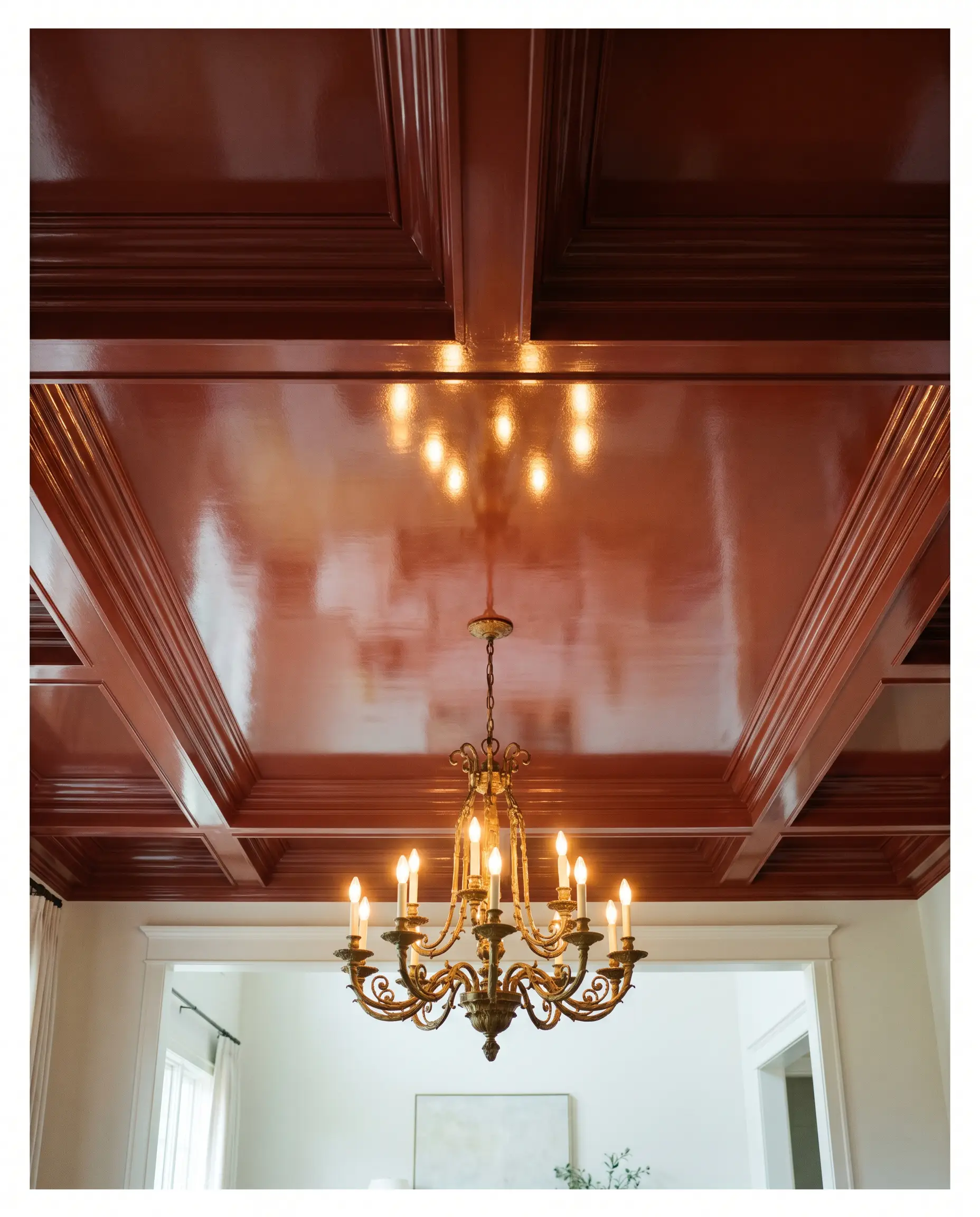 A central unlacquered bronze chandelier bounces light off a brilliant, reflective high-gloss woodwork finish on a dining room tray ceiling painted in a deep auburn cast.