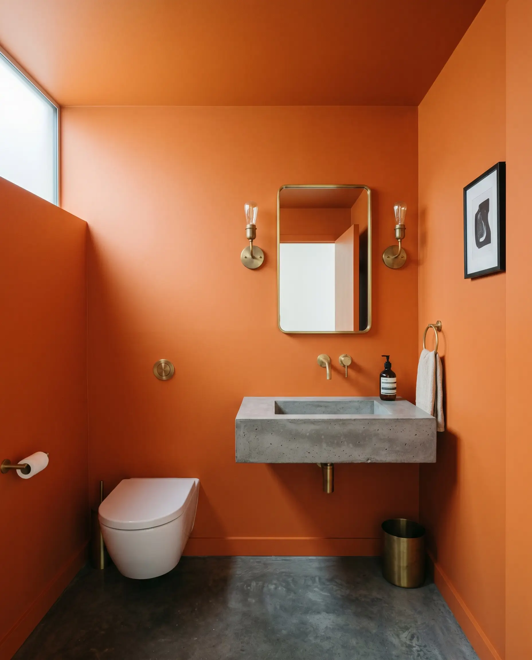 A floating poured concrete sink and unlacquered brass wall sconces highlight a small contemporary powder room painted seamlessly in Benjamin Moore Electric Orange (Hex #F0701A).