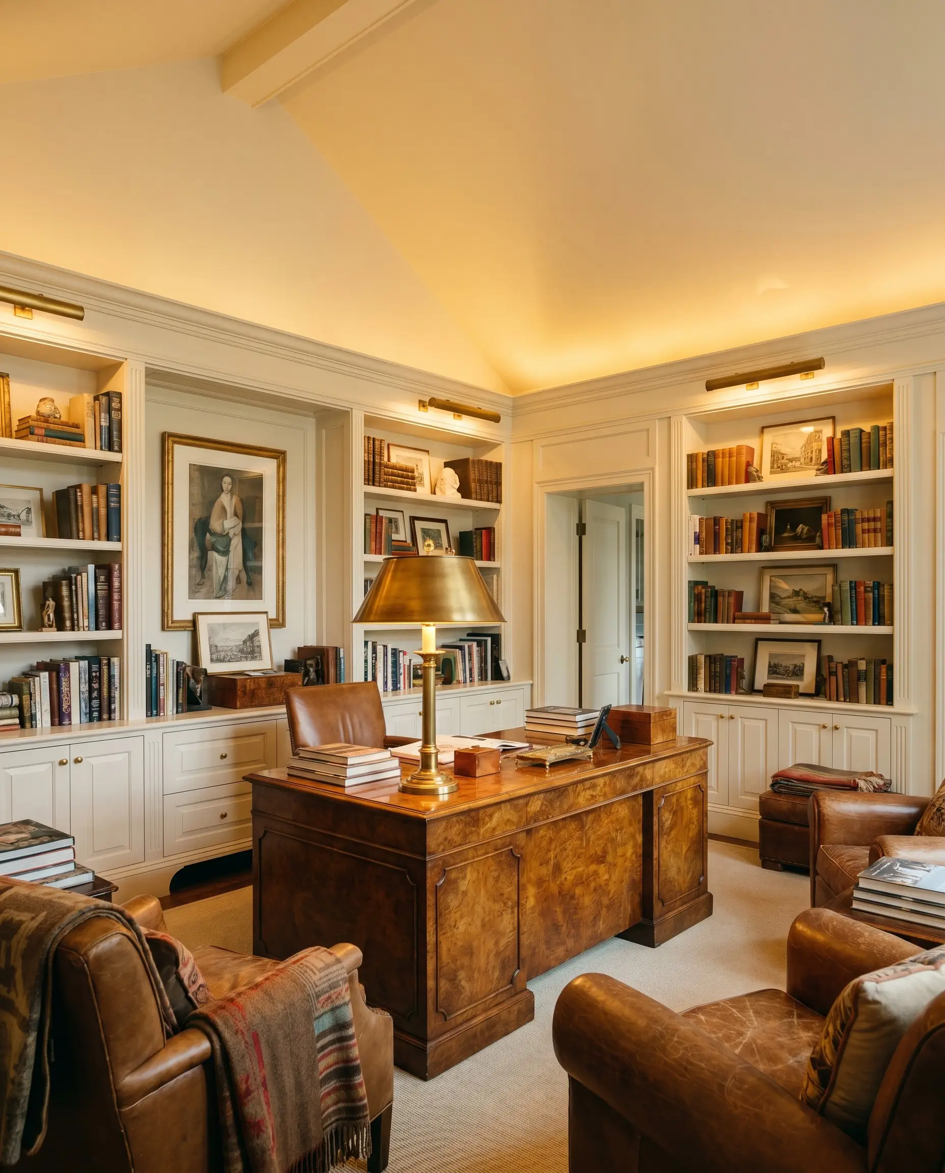A statement brass reading lamp illuminates a highly polished burled wood desk surrounded by seamless warm off-white built-in bookcases and traditional interiors.