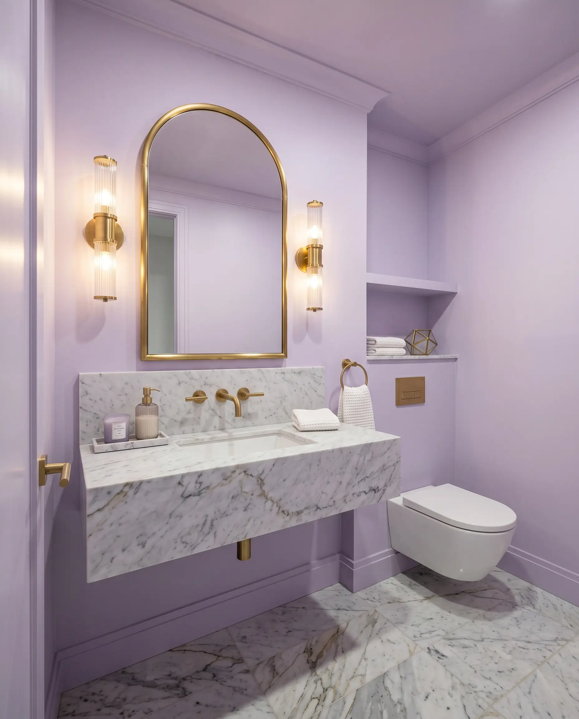 A windowless guest bathroom is completely enveloped in Benjamin Moore Lily Lavender (Hex #DBCEE8), featuring honed Carrara marble floor tiles and unlacquered brass wall sconces.