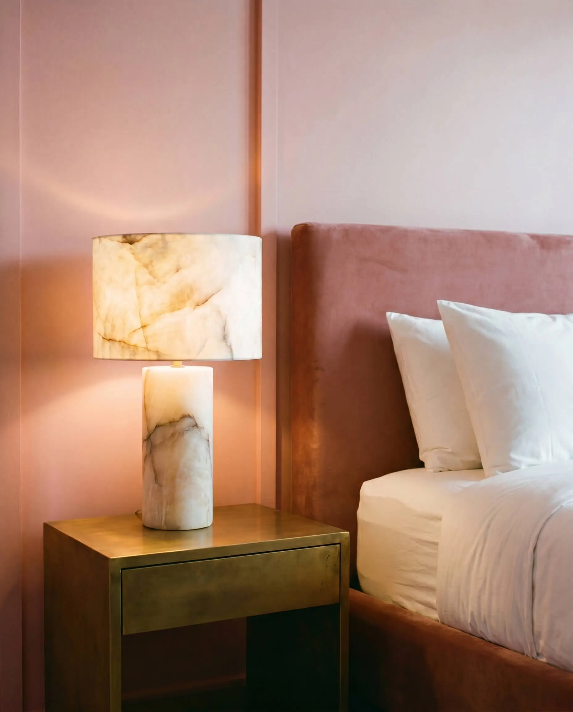 A carved alabaster stone table lamp glows on an unlacquered brass nightstand beside a dusty rose velvet bed in a luxury pink girl bedroom design.