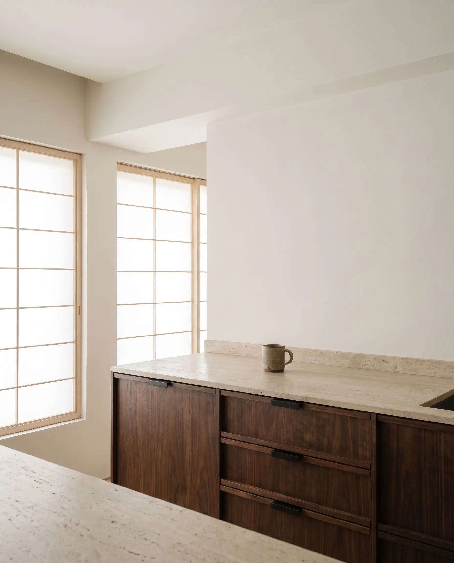 Deep American walnut base cabinets anchor a light leathered travertine countertop styled with a single handmade stoneware mug against bare warm white walls.