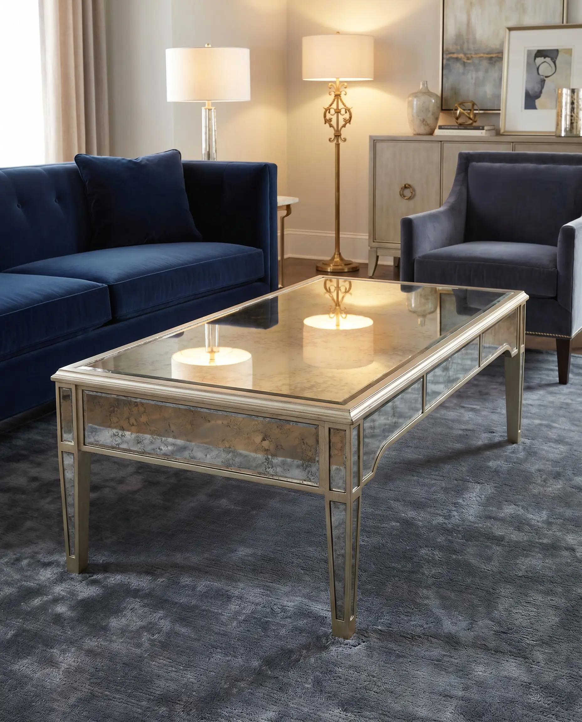 Champagne silver-leafed wood frames antique mirrored panels on a glamorous coffee table resting on a deep-pile slate gray viscose rug beside a tailored navy couch.