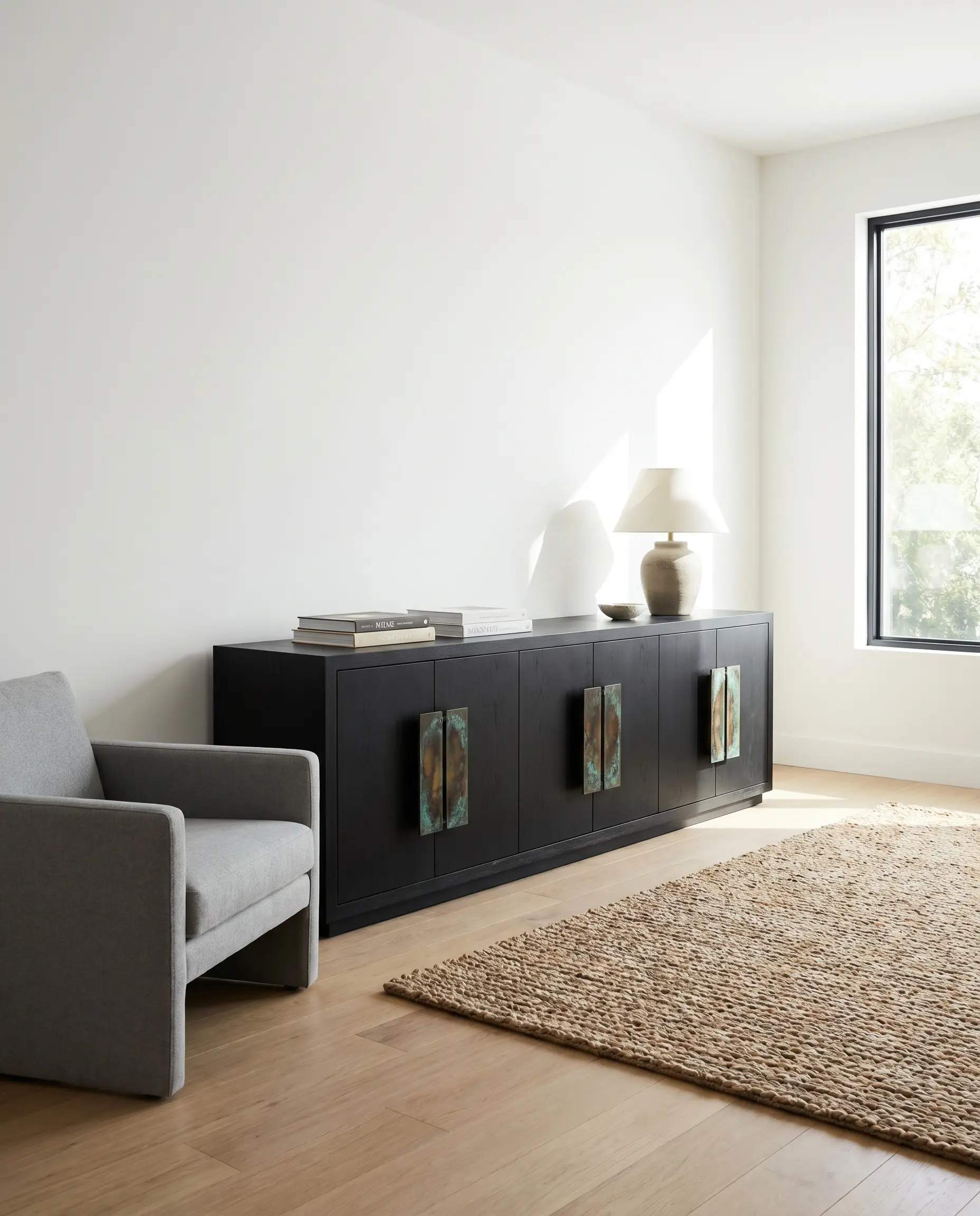Geometric unlacquered brass handles with stepped profiles adorn the doors of a matte modern media console beside a simple chair and textured rug.