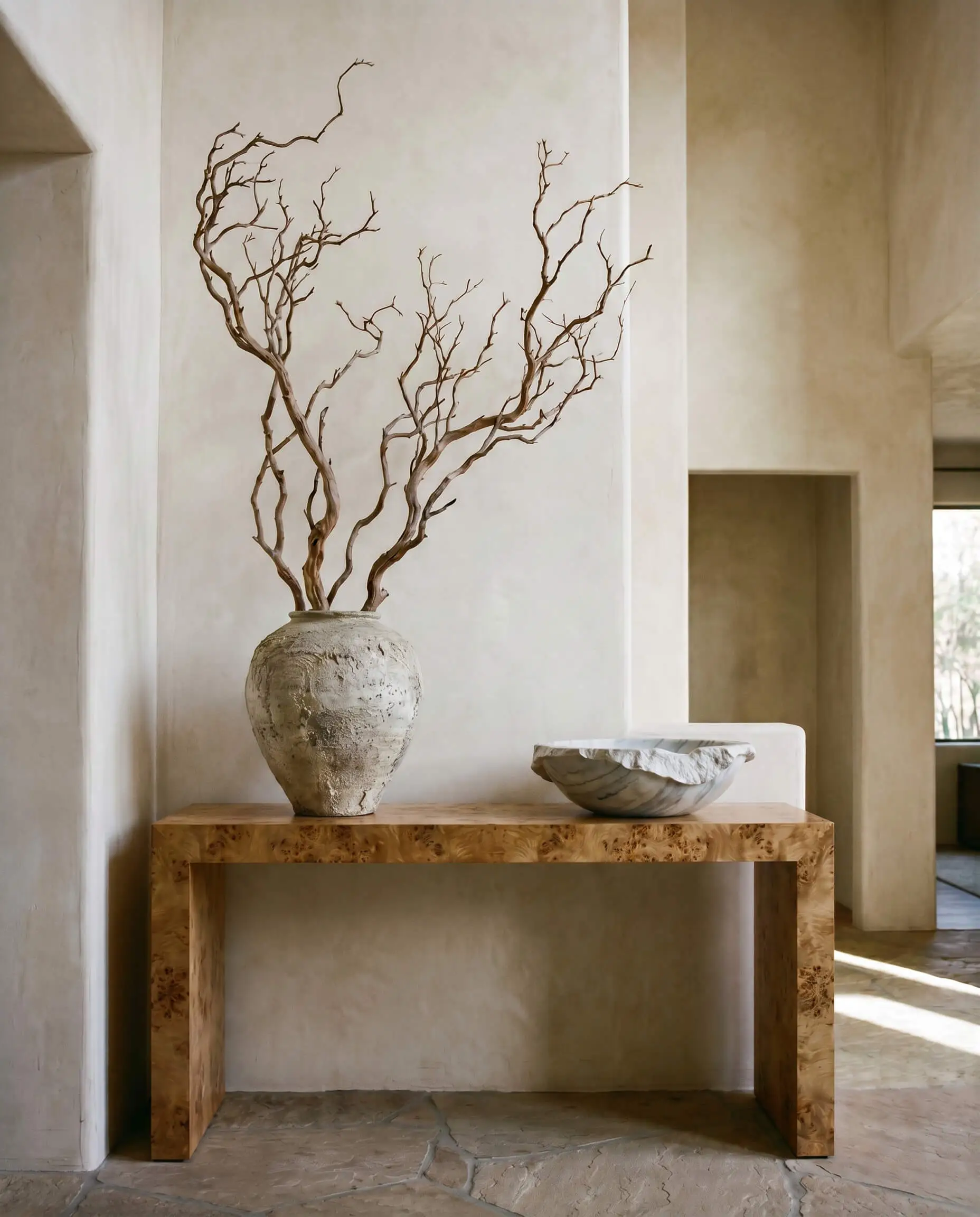 A textured wabi-sabi ceramic vessel holding structural dried manzanita branches creates dynamic asymmetry alongside a low heavy marble bowl on a burl foyer console.