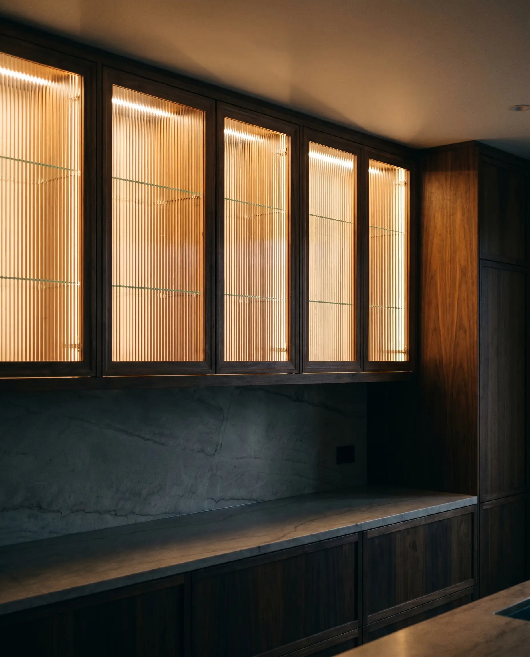 Hidden 3000K LED tape illuminates fluted glass upper cabinets framed in rich wood above a dimly lit marble countertop.