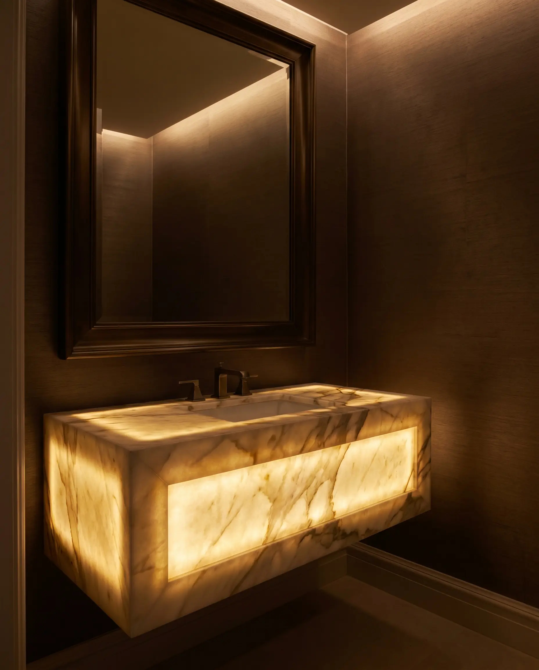 Integrated warm LED panels illuminate a translucent calacatta marble powder room vanity with a deep floating mitered apron beneath dark bronze mirrors.