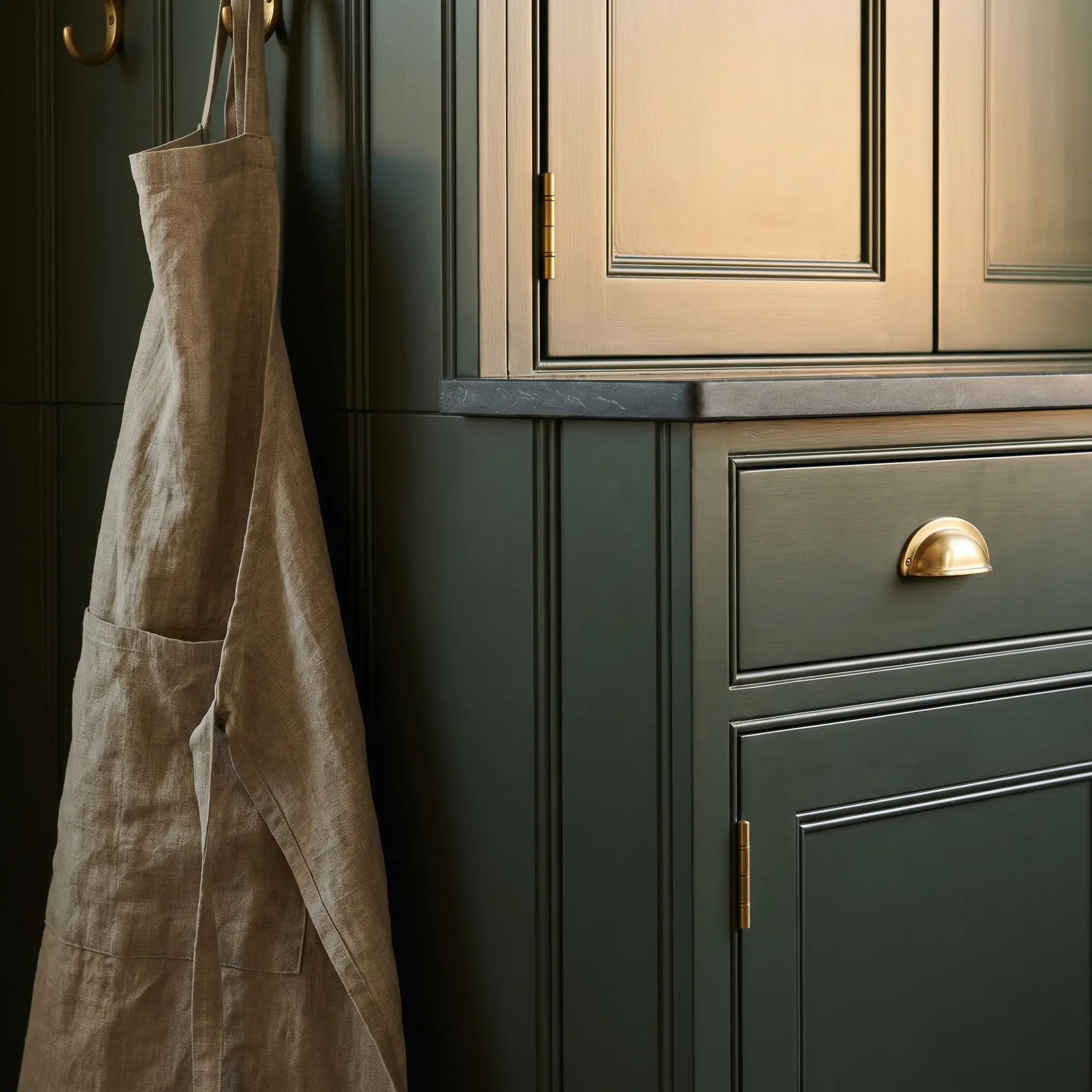 Custom beaded inset face frames on Farrow and Ball Studio Green kitchen cabinetry feature exposed barrel hinges in unlacquered brass beneath an oiled stone surface.