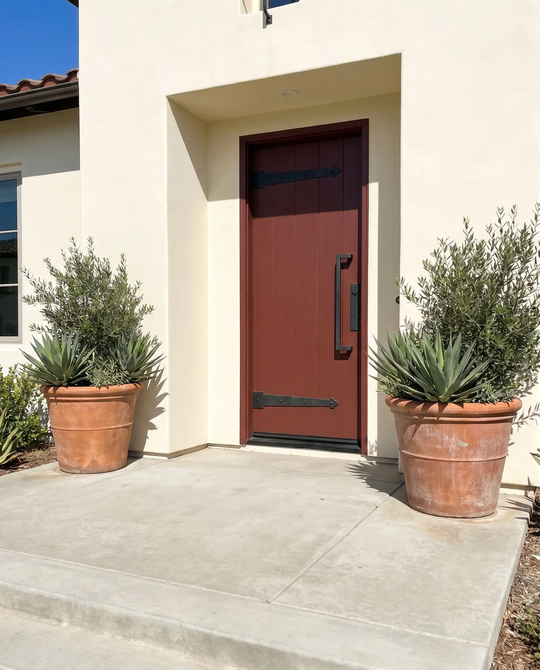 A solid wood exterior door painted an earthy crimson features heavy blackened steel hardware, flanked by oversized terracotta planters on a raw poured concrete porch.