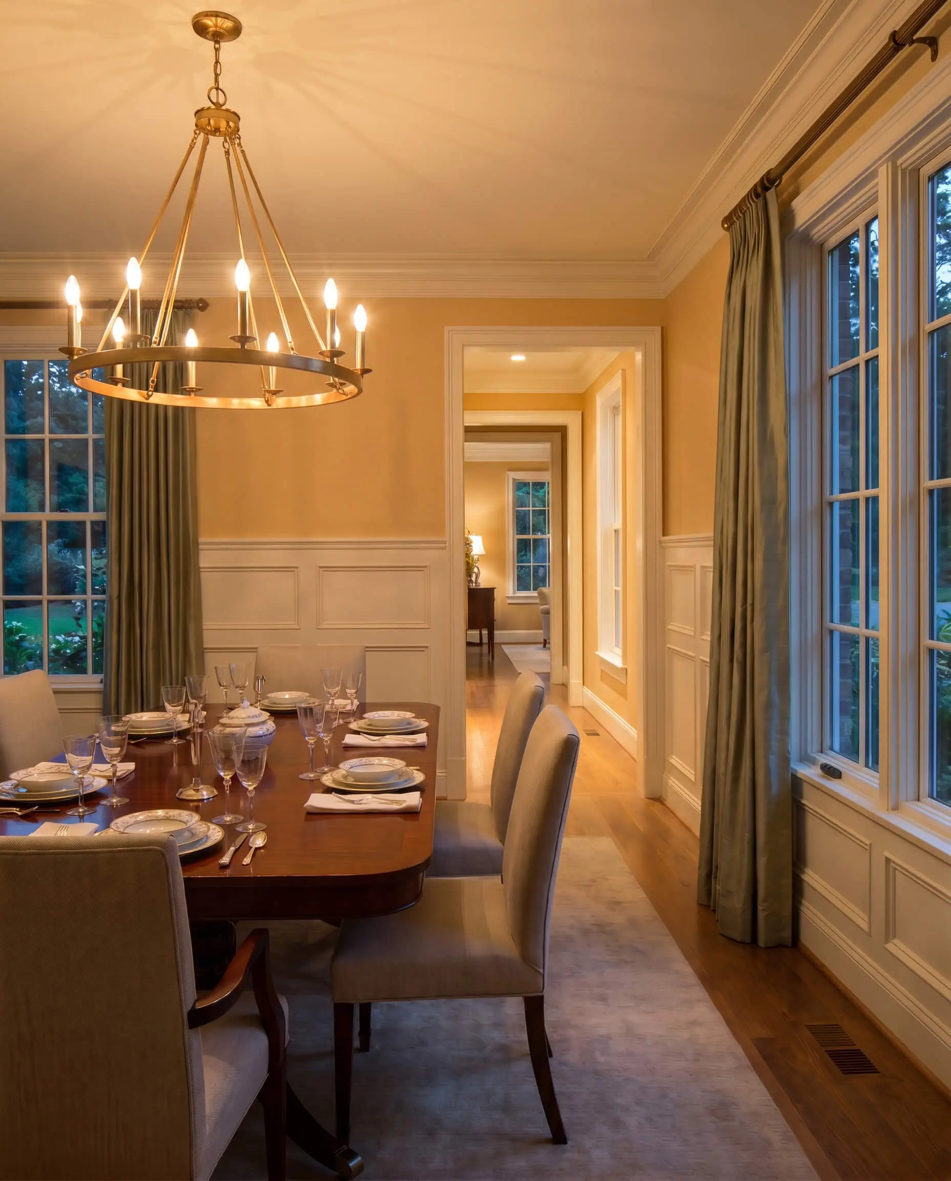 An unlacquered brass chandelier casts a warm glow over a polished mahogany dining table, framed by loden green drapery and golden tan upper walls.