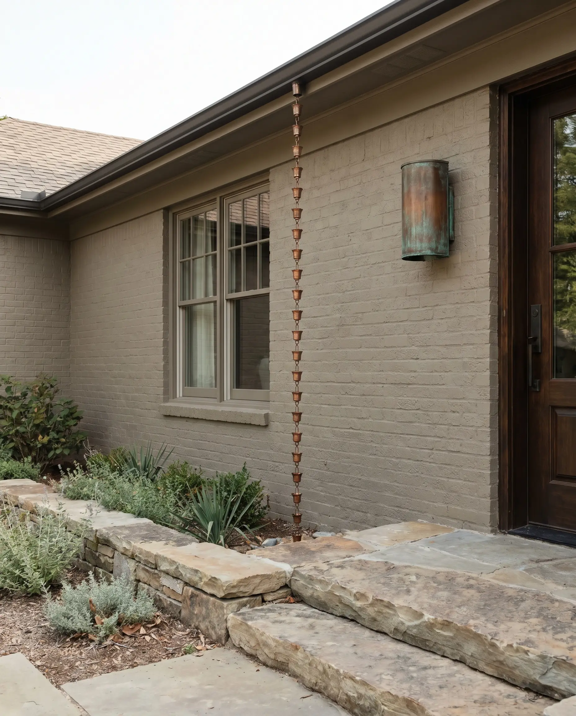 Benjamin Moore Revere Pewter coats a brick exterior accented by a raw copper rain chain, unlacquered copper wall sconces, and natural stone hardscaping.