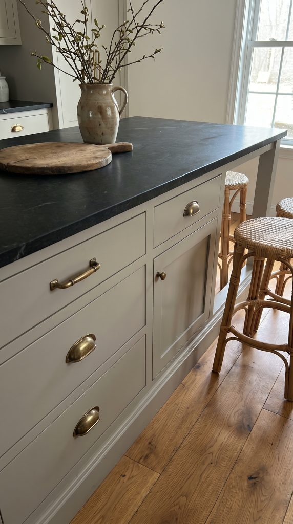 A warm gray kitchen island topped with a thick slab of honed soapstone features heavy aged brass cup pulls and woven counter stools.