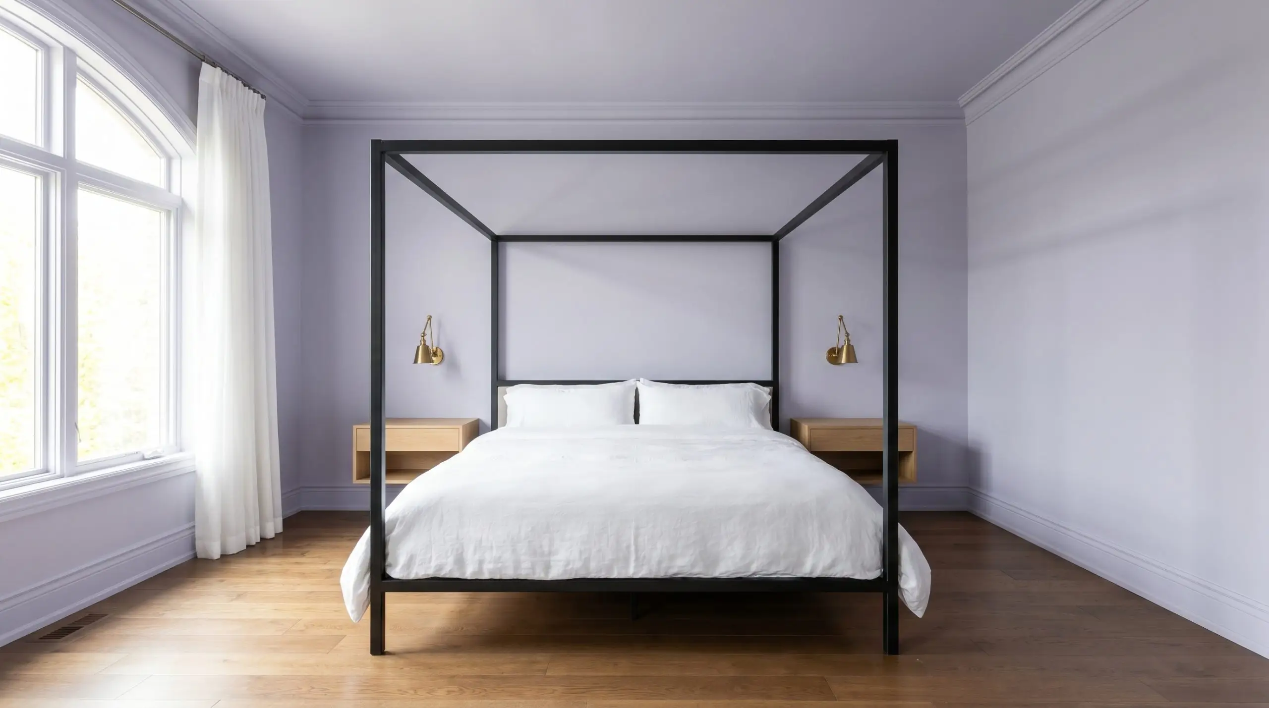 A sharp blackened steel canopy bed dressed in white washed linen anchors a serene guest bedroom surrounded by cool-toned neutral walls and brass sconces.