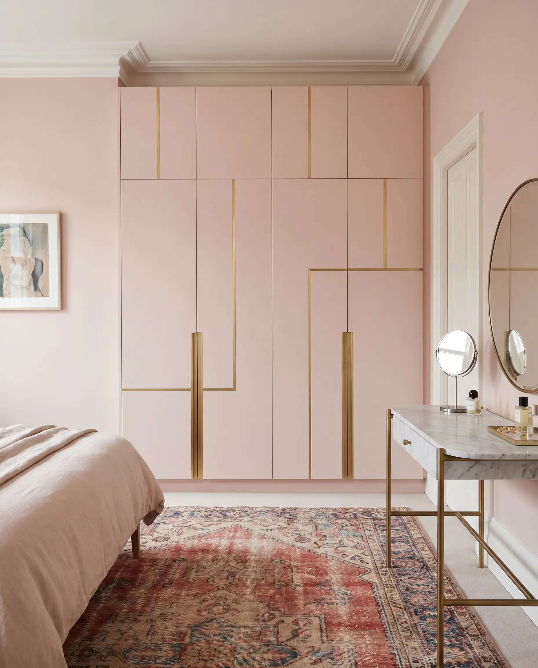 Flush-mount floor-to-ceiling millwork wardrobes painted blush pink feature brushed brass inlay strips beside a marble-topped vanity and a vintage Turkish rug.