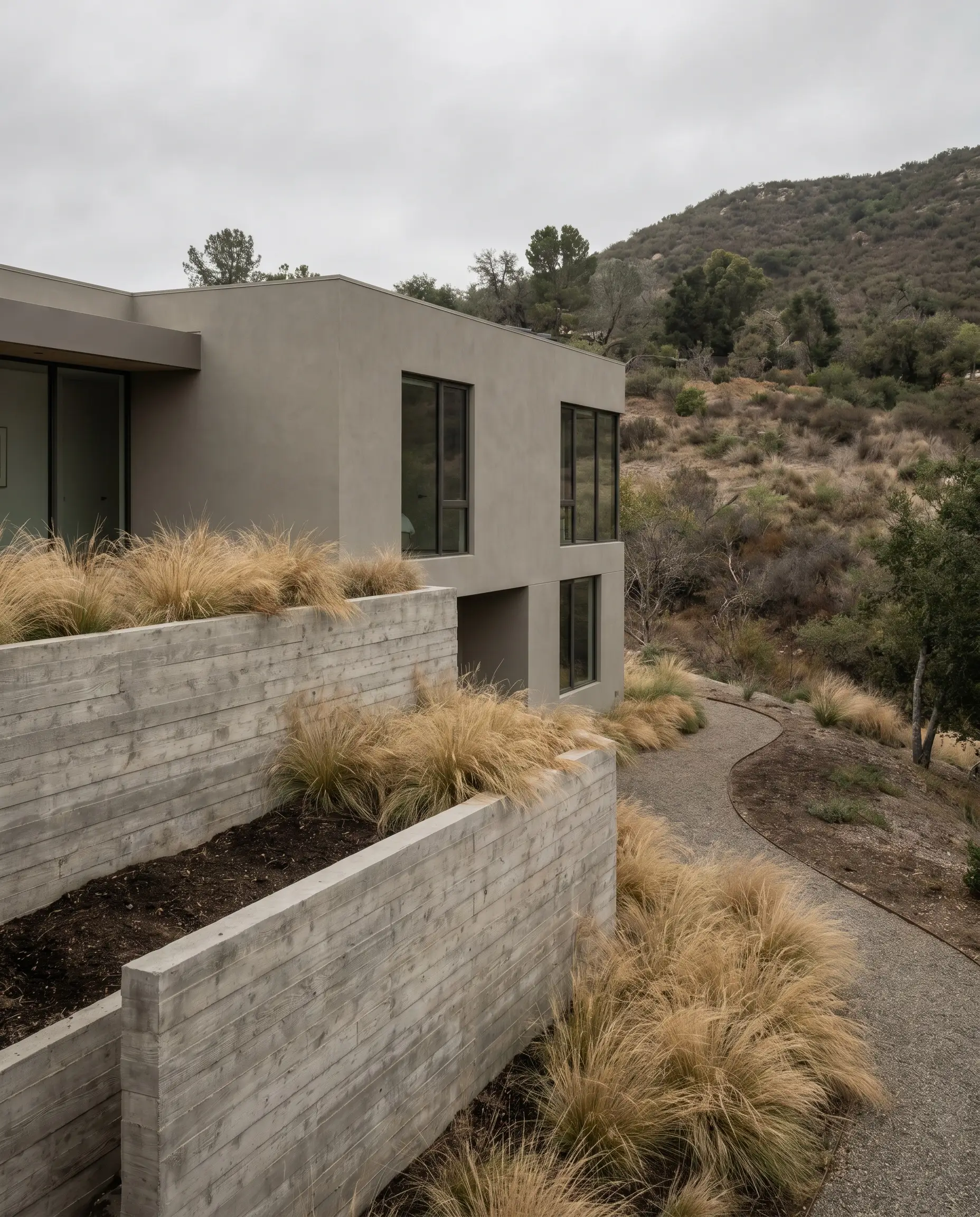 Raw board-formed concrete retaining walls anchor a smooth greige-painted upper level surrounded by ornamental grasses spilling over the textured concrete foundation.