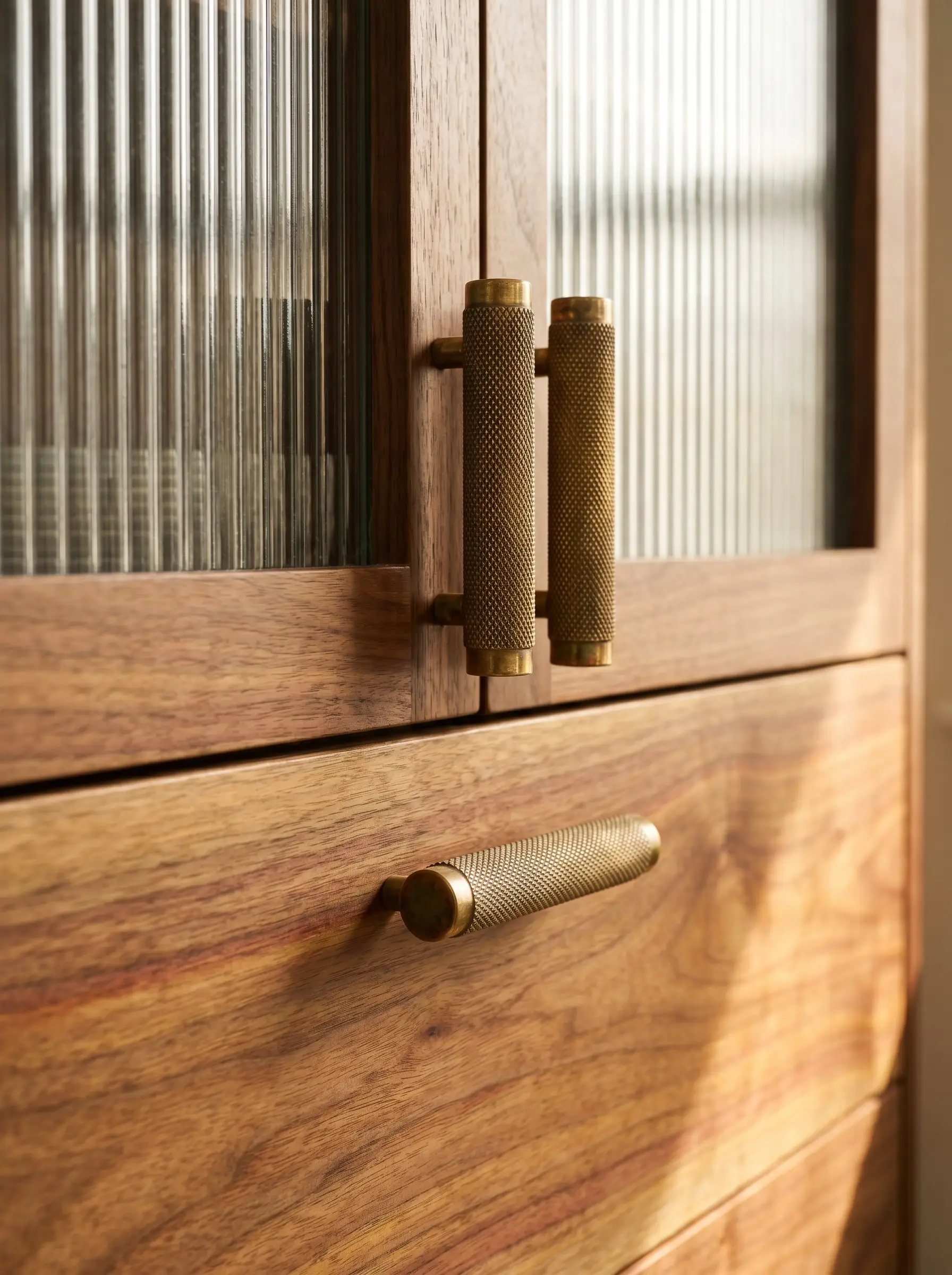 Solid unlacquered brass cylinder pulls with diamond knurling bridge a flat-slab wood base drawer and a ribbed glass upper door.