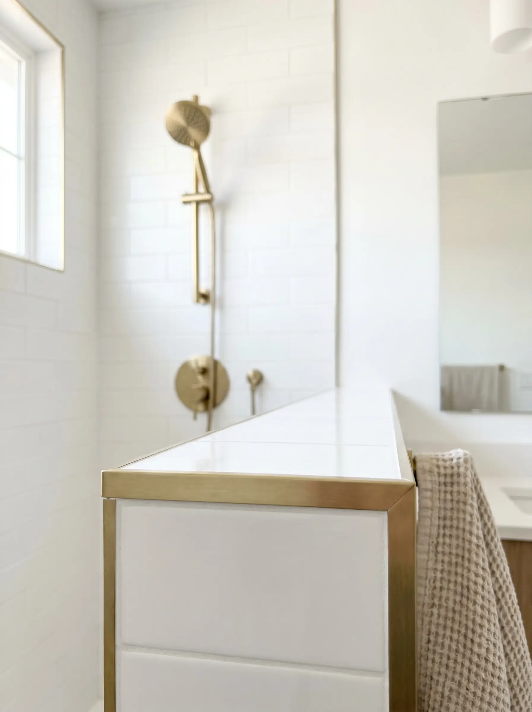 A brushed brass Schluter profile frames the outside corner of a white tiled shower curb, matching the nearby brass showerhead and temperature valve.
