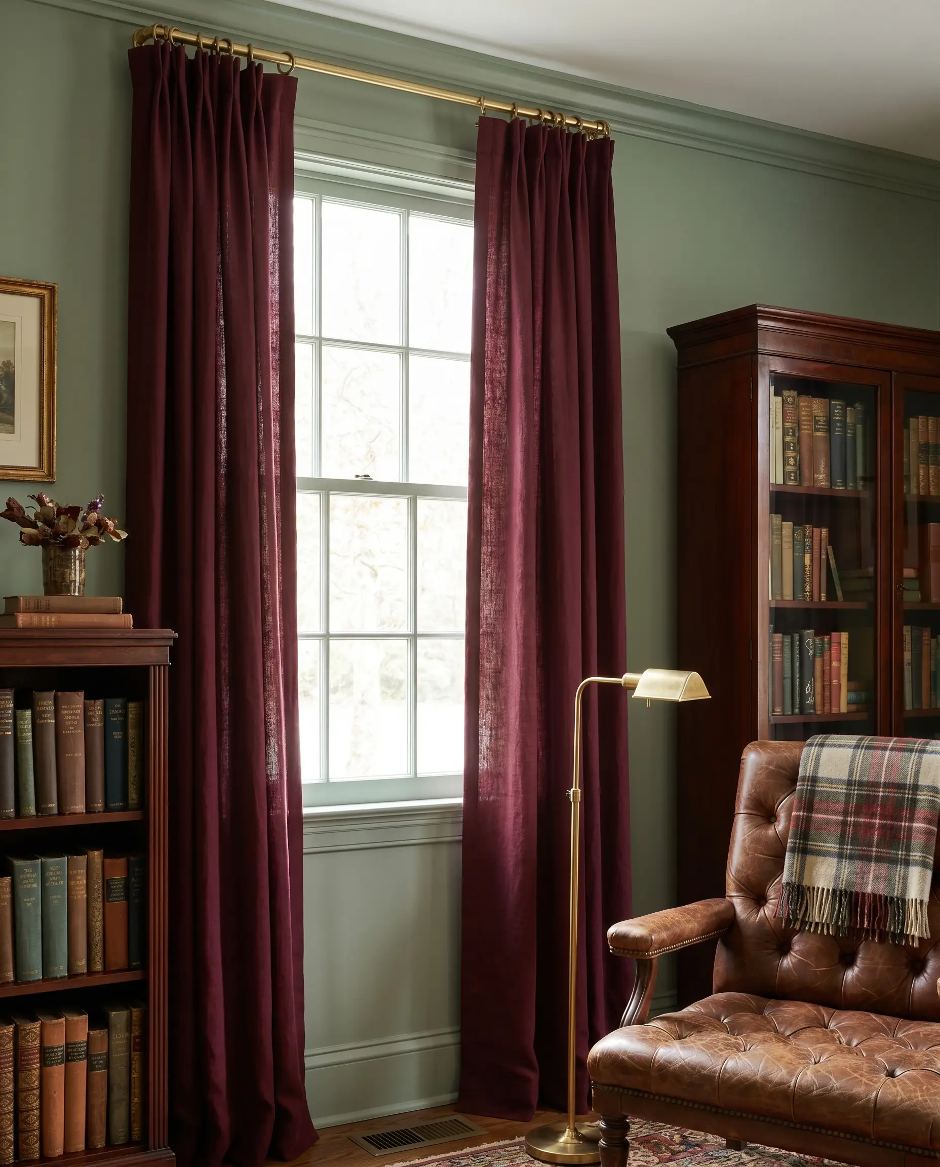 Deep burgundy light-filtering panels frame curtains for sage green walls alongside a mahogany bookshelf and a tufted reading chair with a plaid throw.