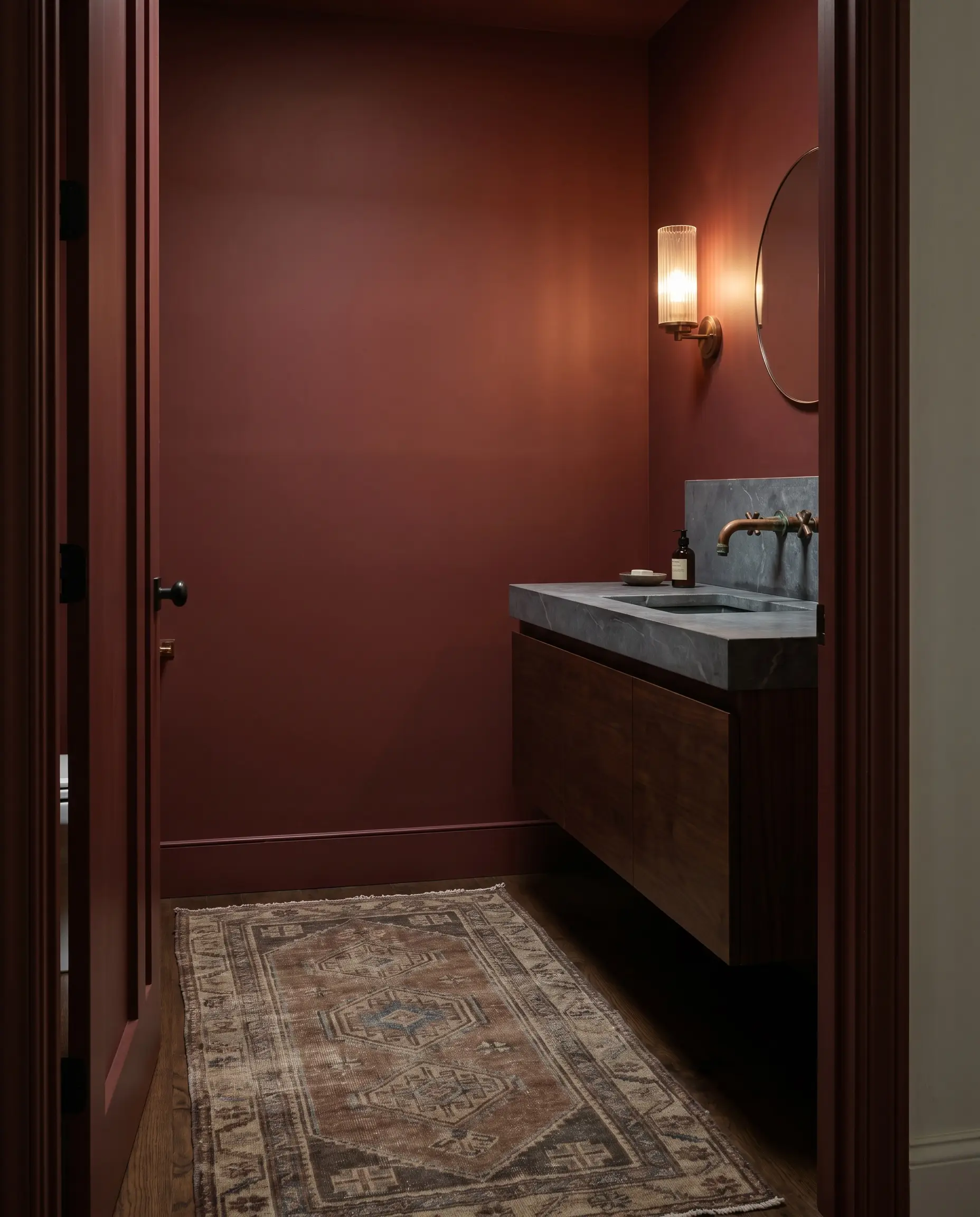 A floating vanity with a honed soapstone countertop and unlacquered copper faucet rests against moody dark red walls alongside a vintage wool runner.