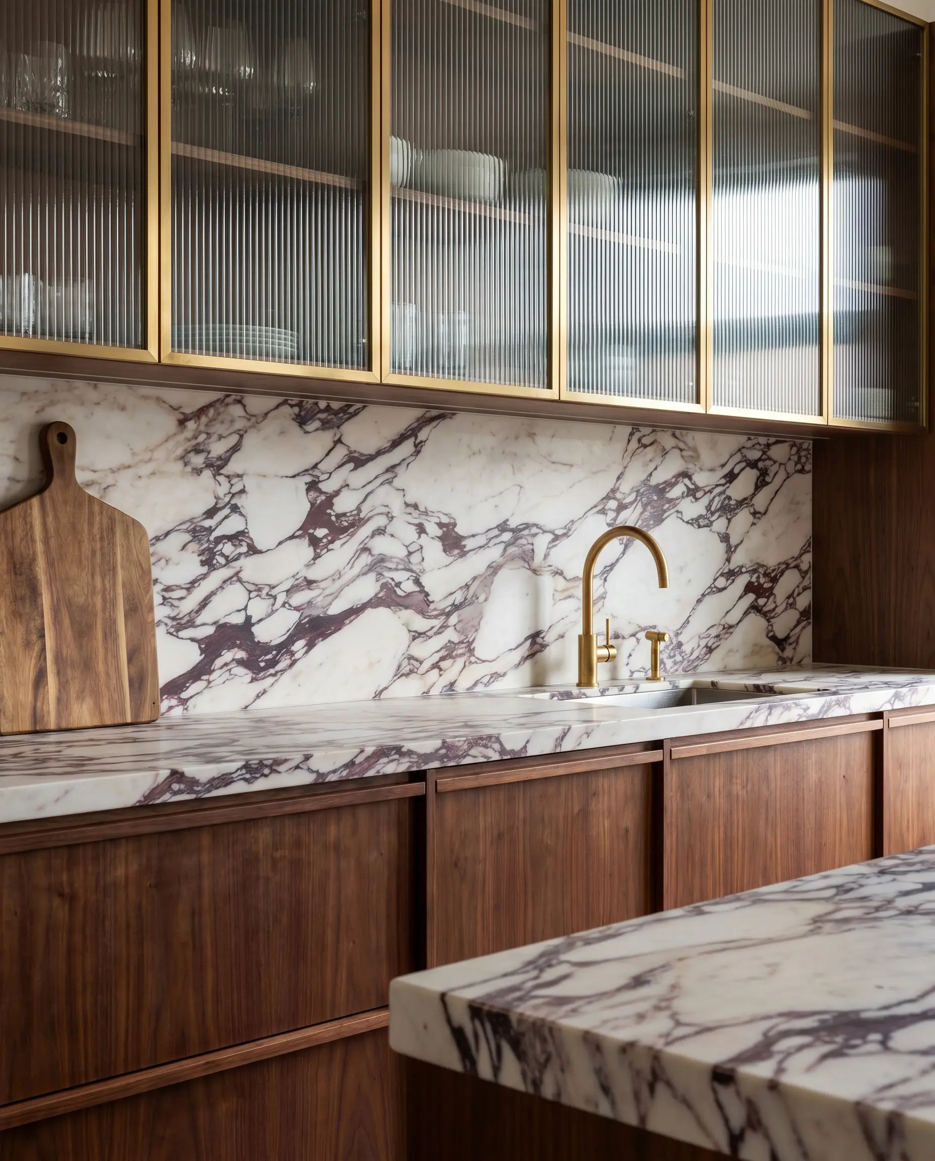 A heavily veined Calacatta Viola marble countertop and continuous slab backsplash separate reeded glass and walnut kitchen cabinet fronts.