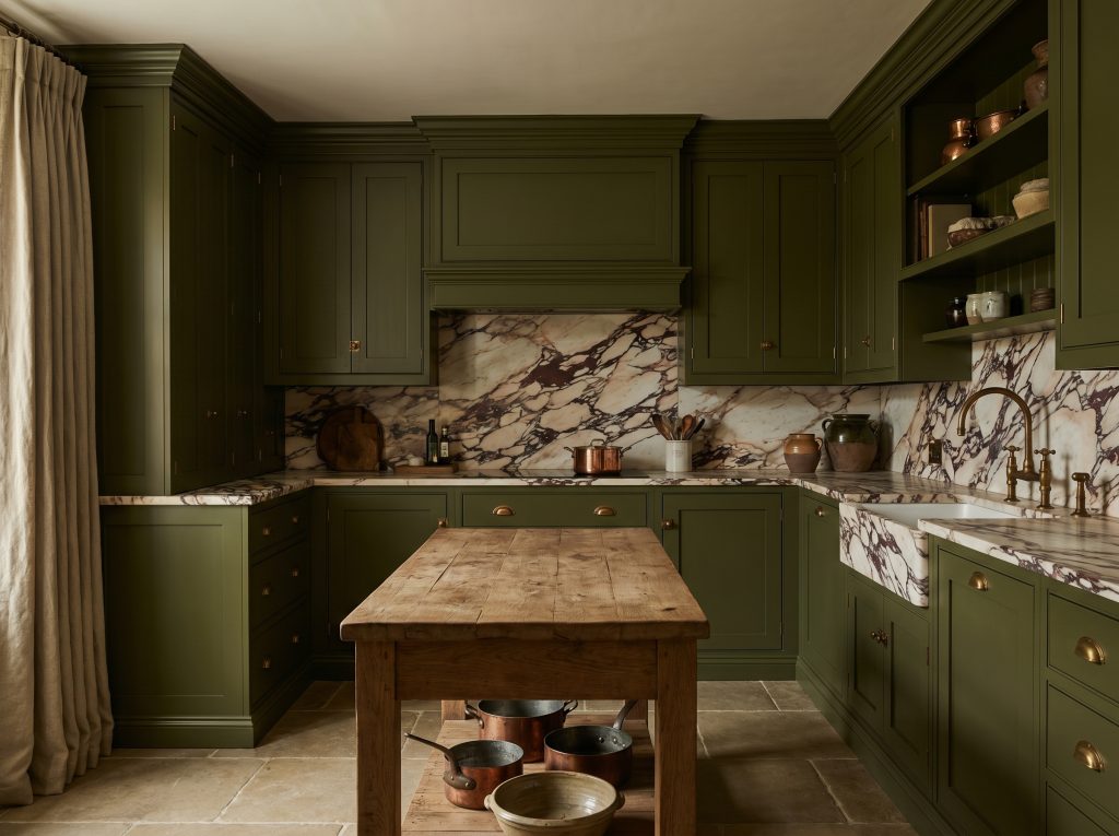 Rich historic olive green cabinetry surrounds dramatic veined marble countertops, unlacquered brass cup pulls, and a rustic wooden prep table.