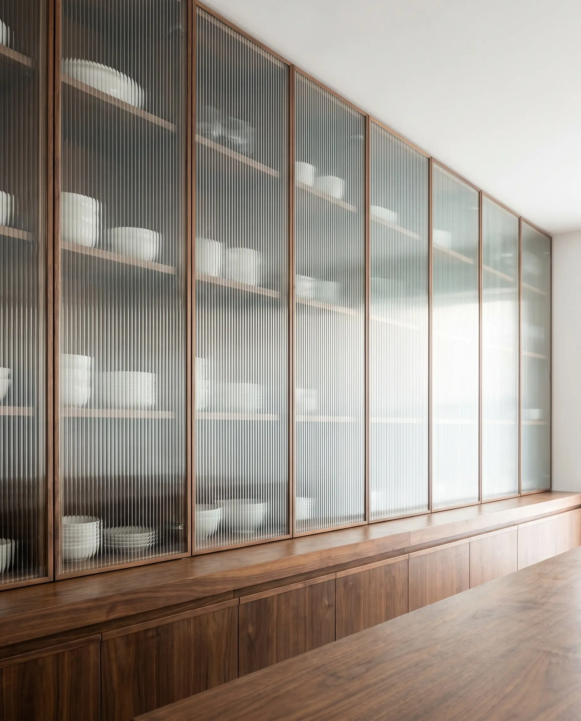 Tall reeded glass and walnut kitchen cabinet fronts stretch to a pure white ceiling above thick wood countertops and base units.