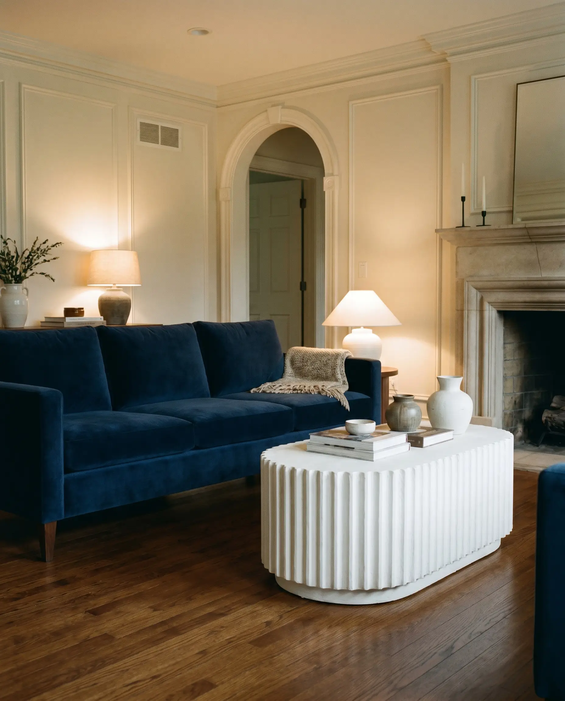 Deep vertical ridges define a chalk white fluted plaster pedestal coffee table placed on warm walnut hardwood flooring opposite a navy blue velvet three-seater sofa.
