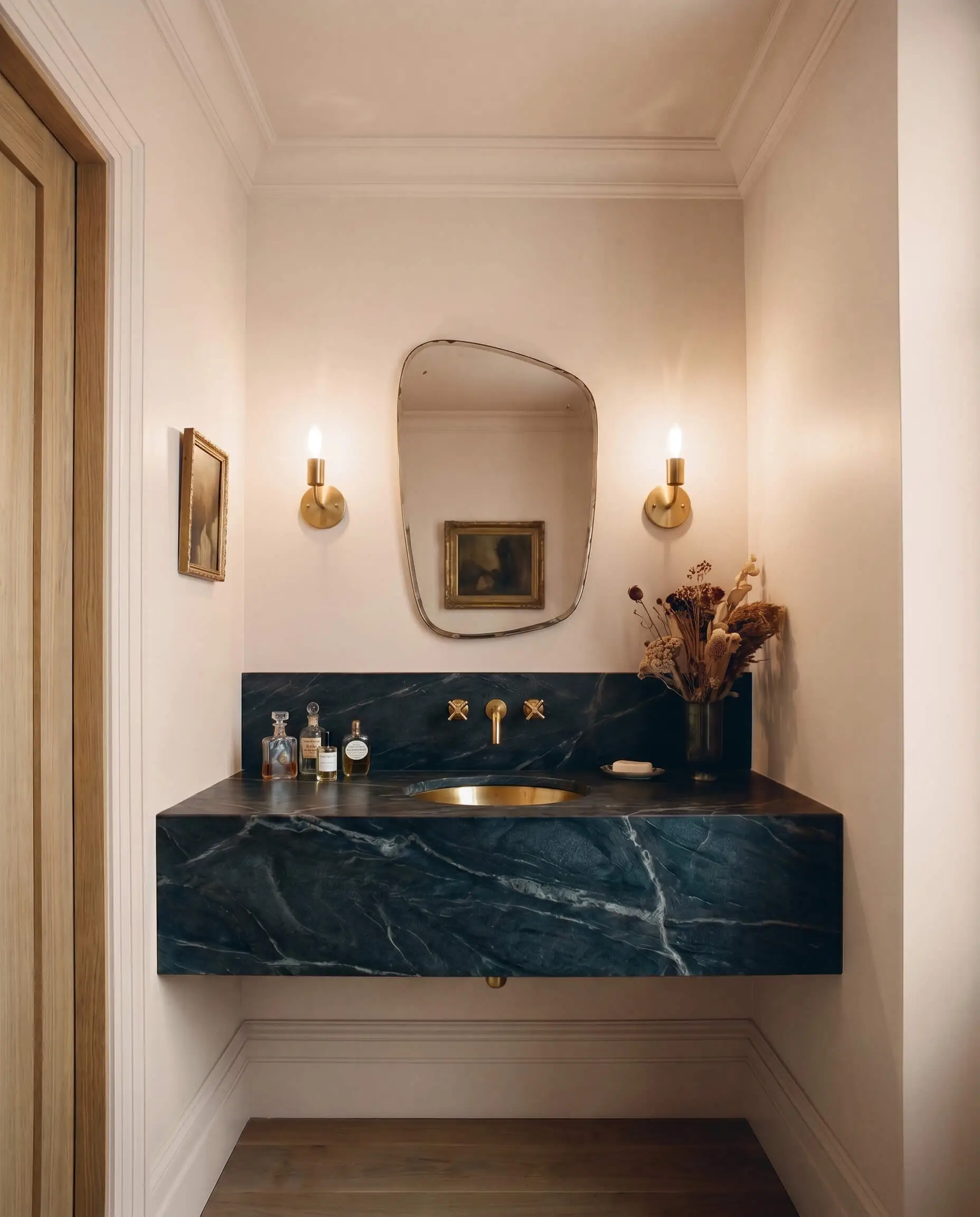 Unlacquered brass wall sconces flank an asymmetrical mirror above a charcoal soapstone vanity in a moody Parisian powder room painted in Benjamin Moore Head Over Heels.