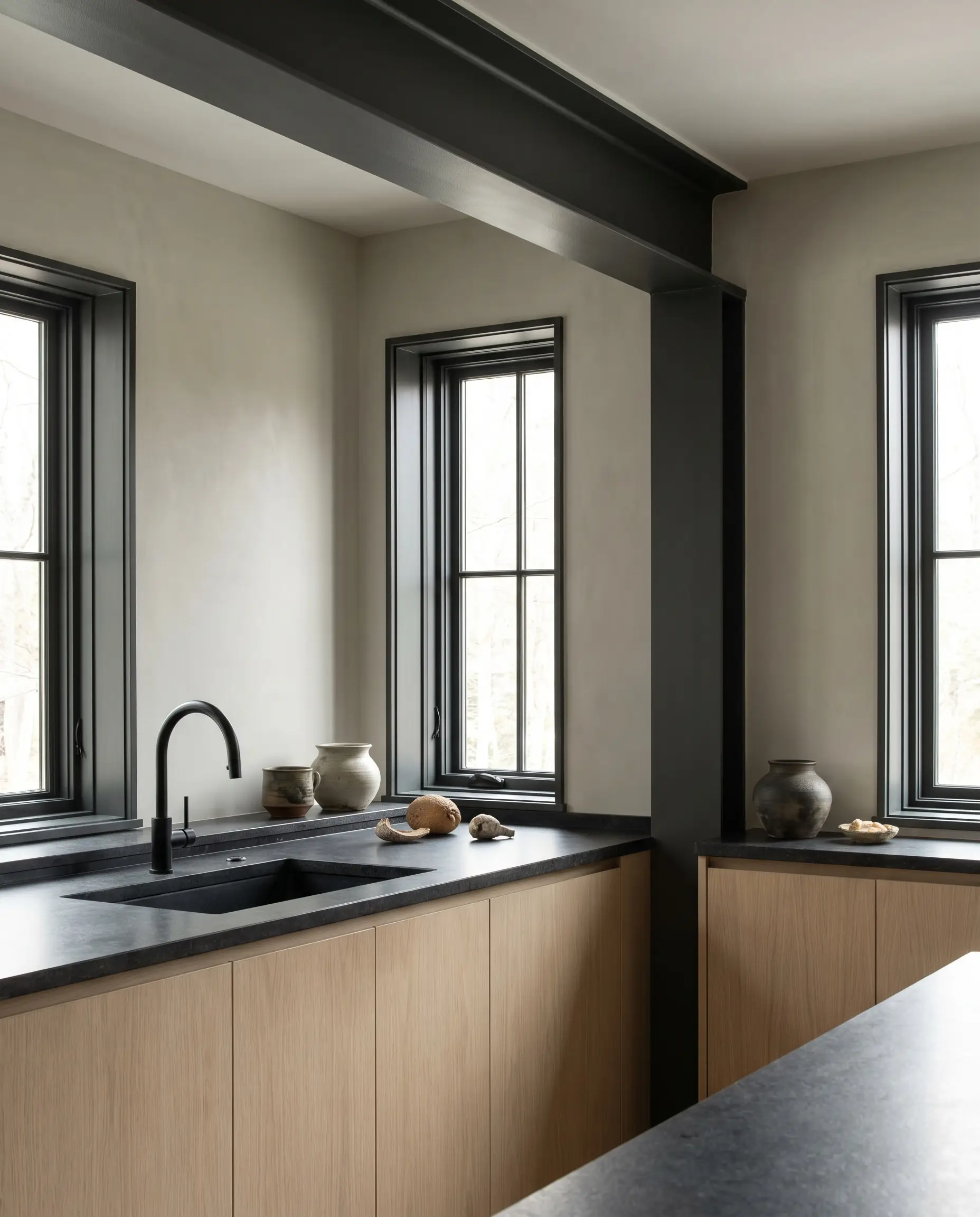 Japandi style kitchens incorporate Benjamin Moore Cheating Heart charcoal accents on window mullions and exposed beams to contrast pale wood cabinetry and greige walls.