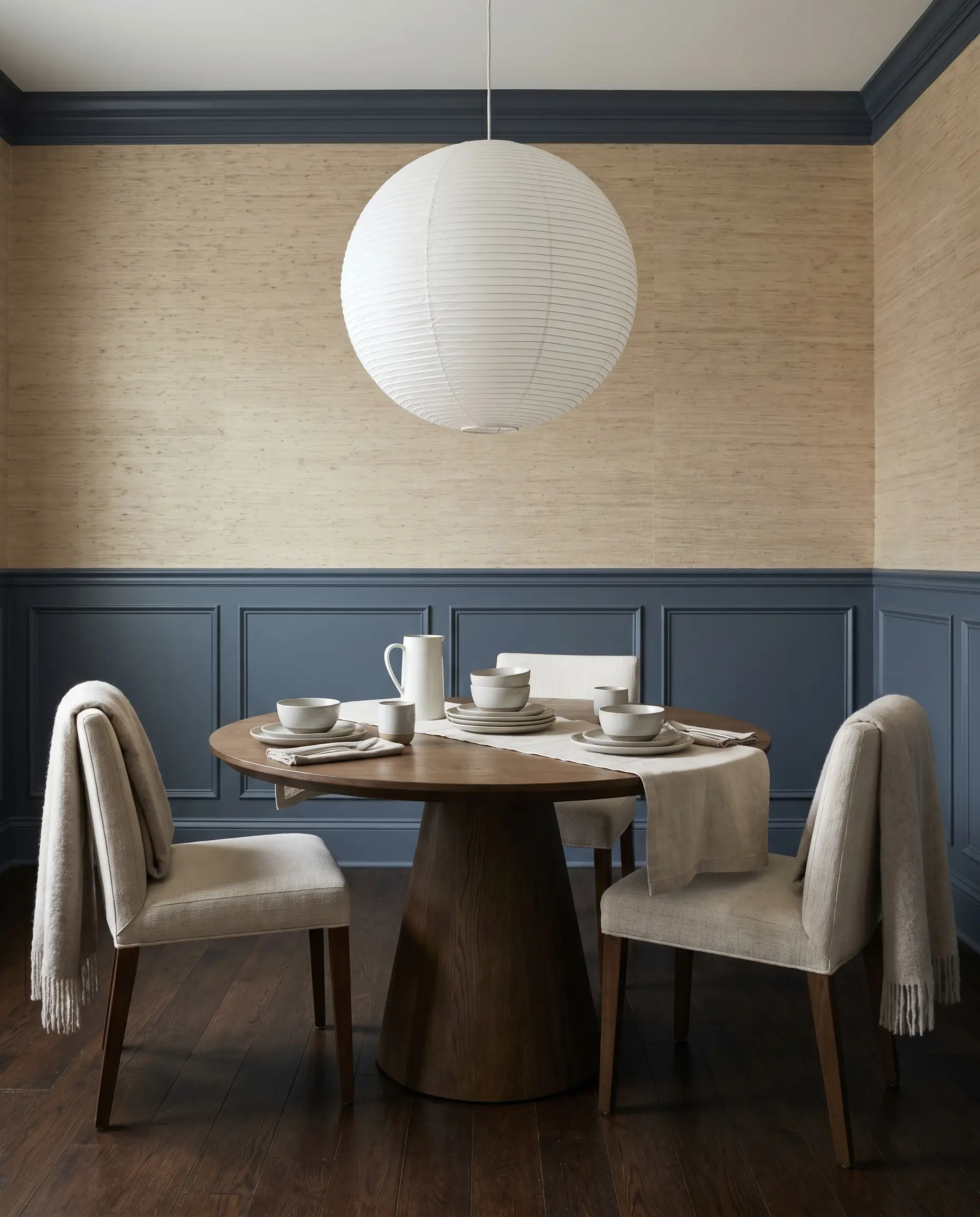 Classic lower wainscoting painted in a moody gray-blue chromatic profile grounds a tailored dining space beneath textured neutral grasscloth wallpaper and an oversized white paper pendant light.