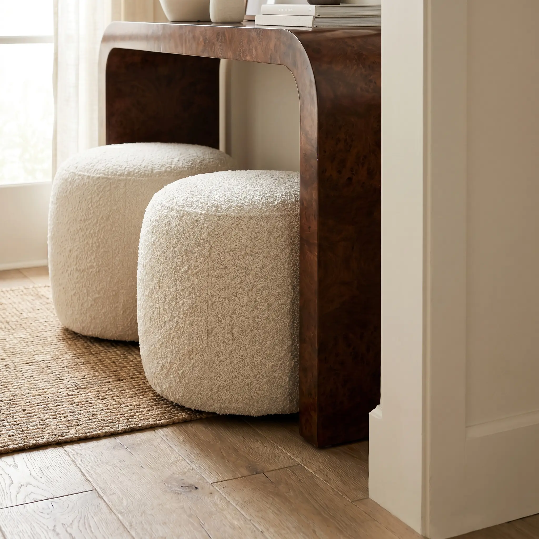 Two rounded heavyweight matte cream boucle ottomans sit neatly tucked beneath a tall, dark brown burl wood waterfall console table.